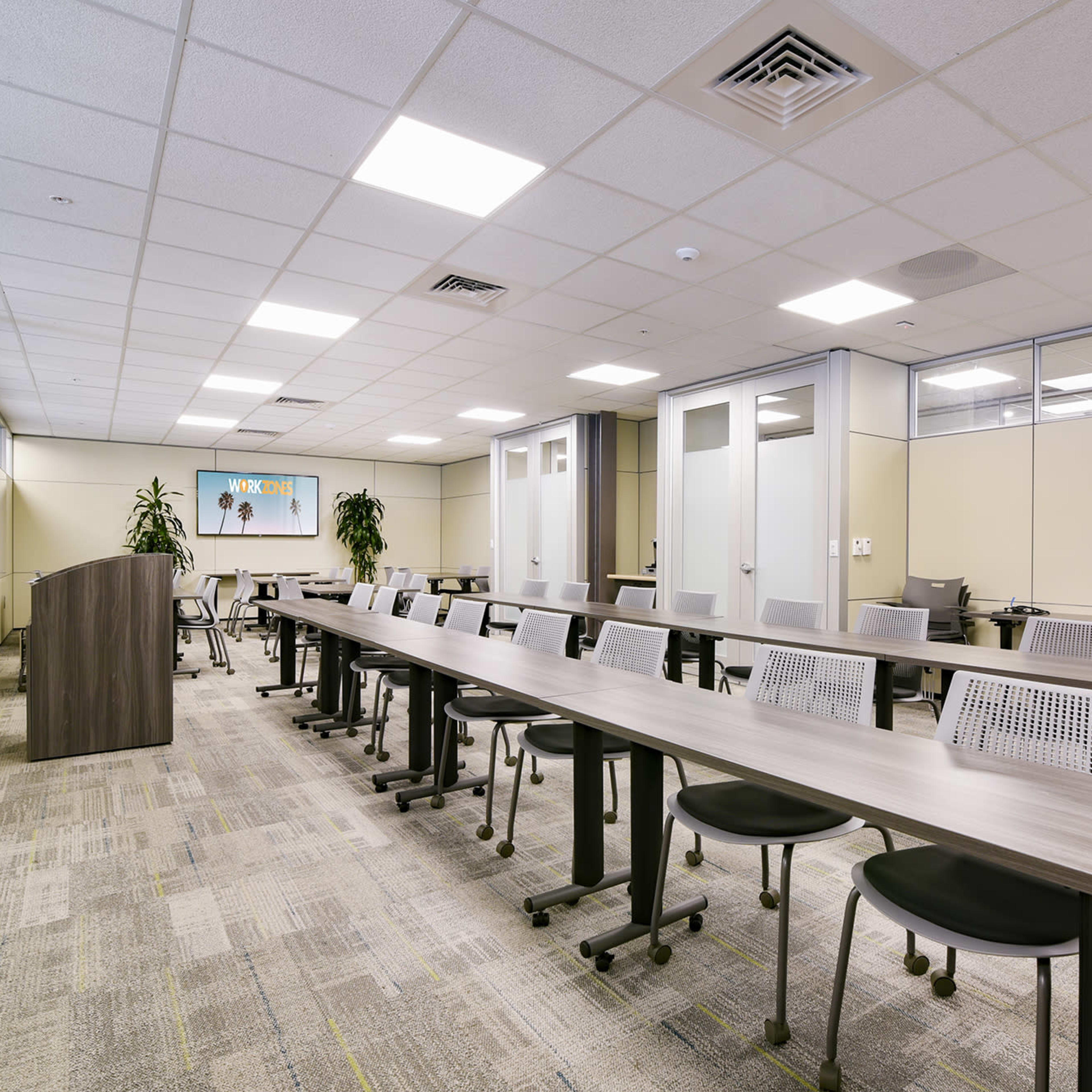 A modern conference room with long tables arranged in a row, accompanied by rolling chairs and plants in the corners.