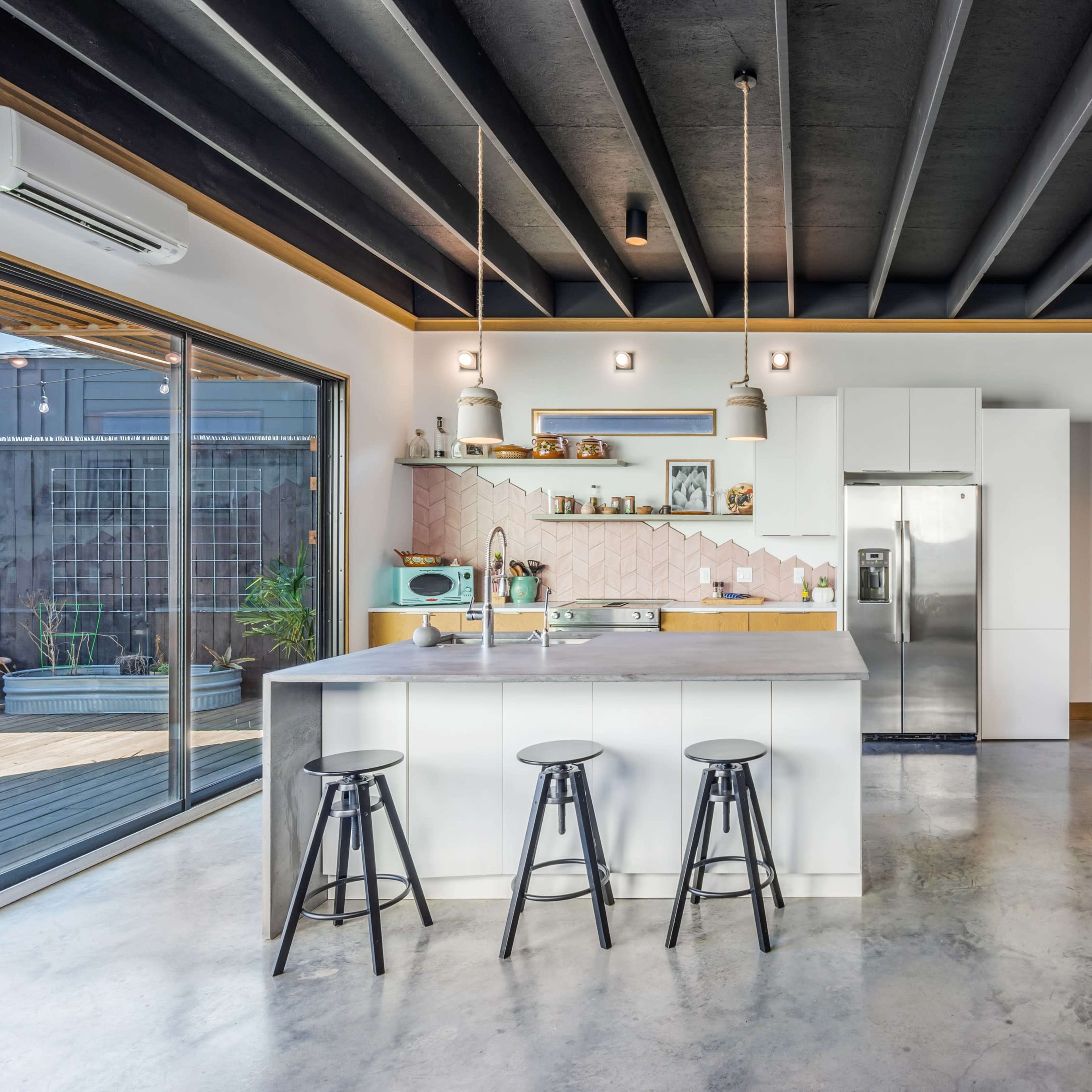 A modern kitchen with a white island and three black stools, alongside a dining area featuring a wooden table and four chairs, illuminated by pendant lights against a backdrop of exposed beams and a large sliding glass door.