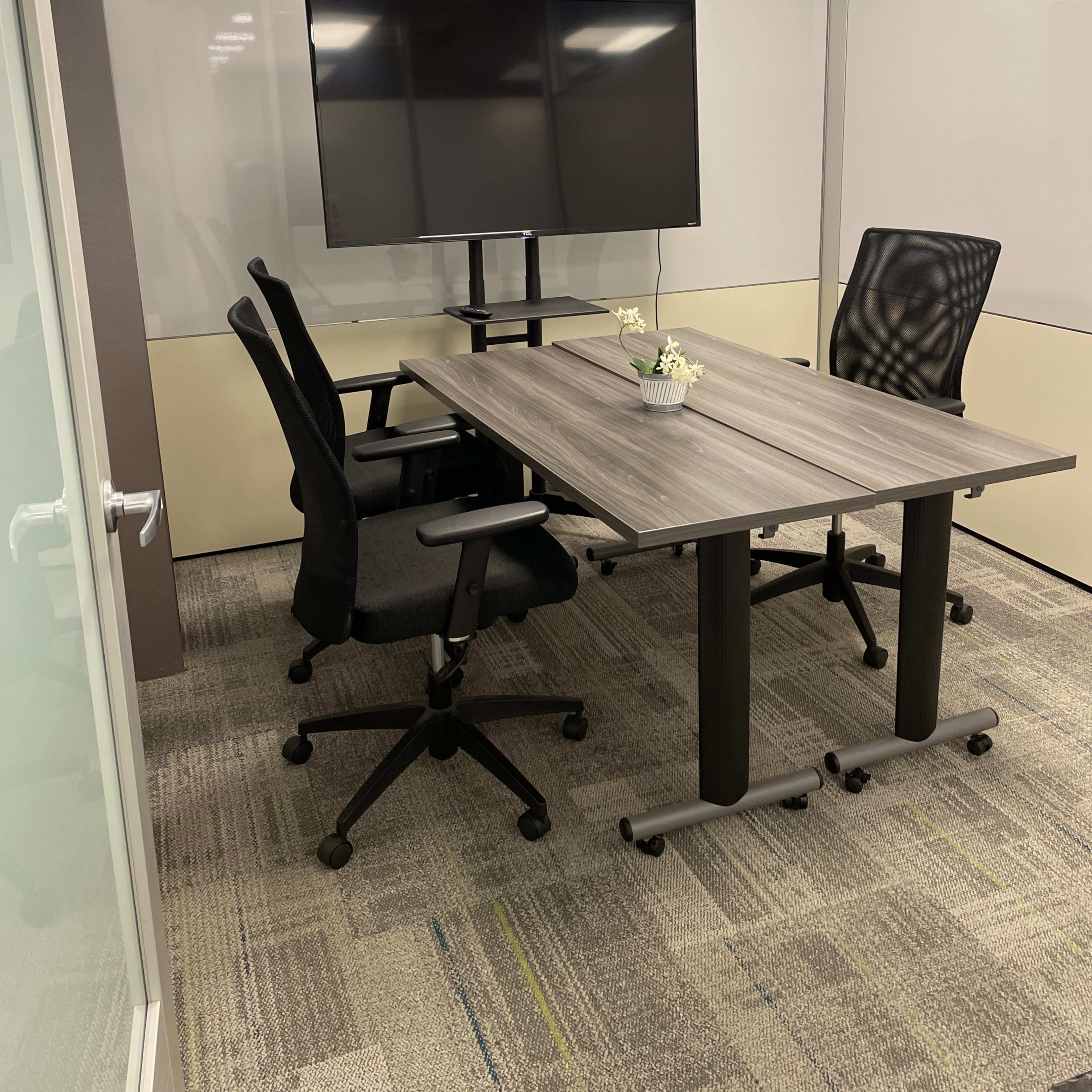 A small meeting room with a wooden table, two office chairs, and a wall-mounted television.