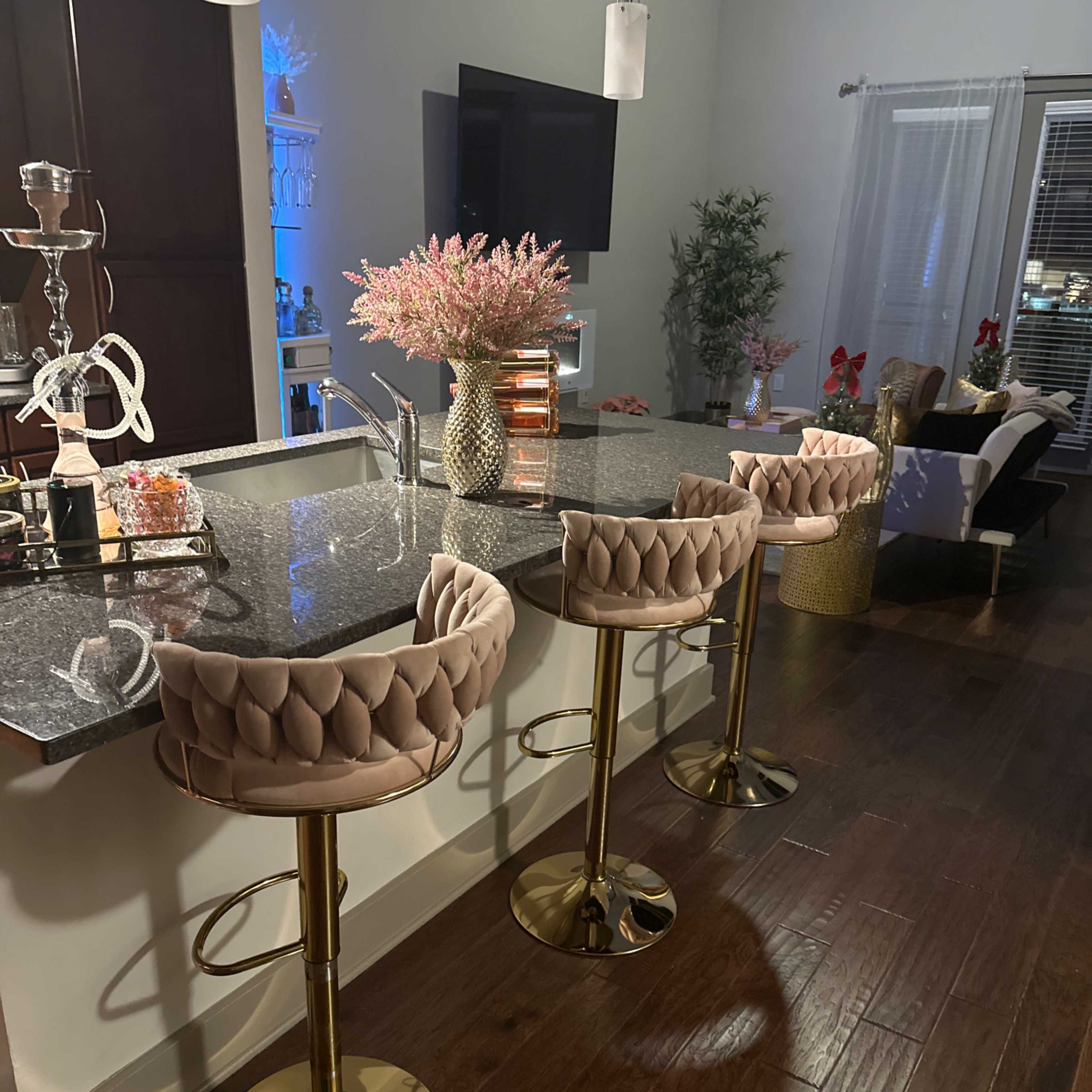 A modern kitchen bar area with three upholstered stools, a marble countertop, a decorative flower arrangement, and a television mounted on the wall.