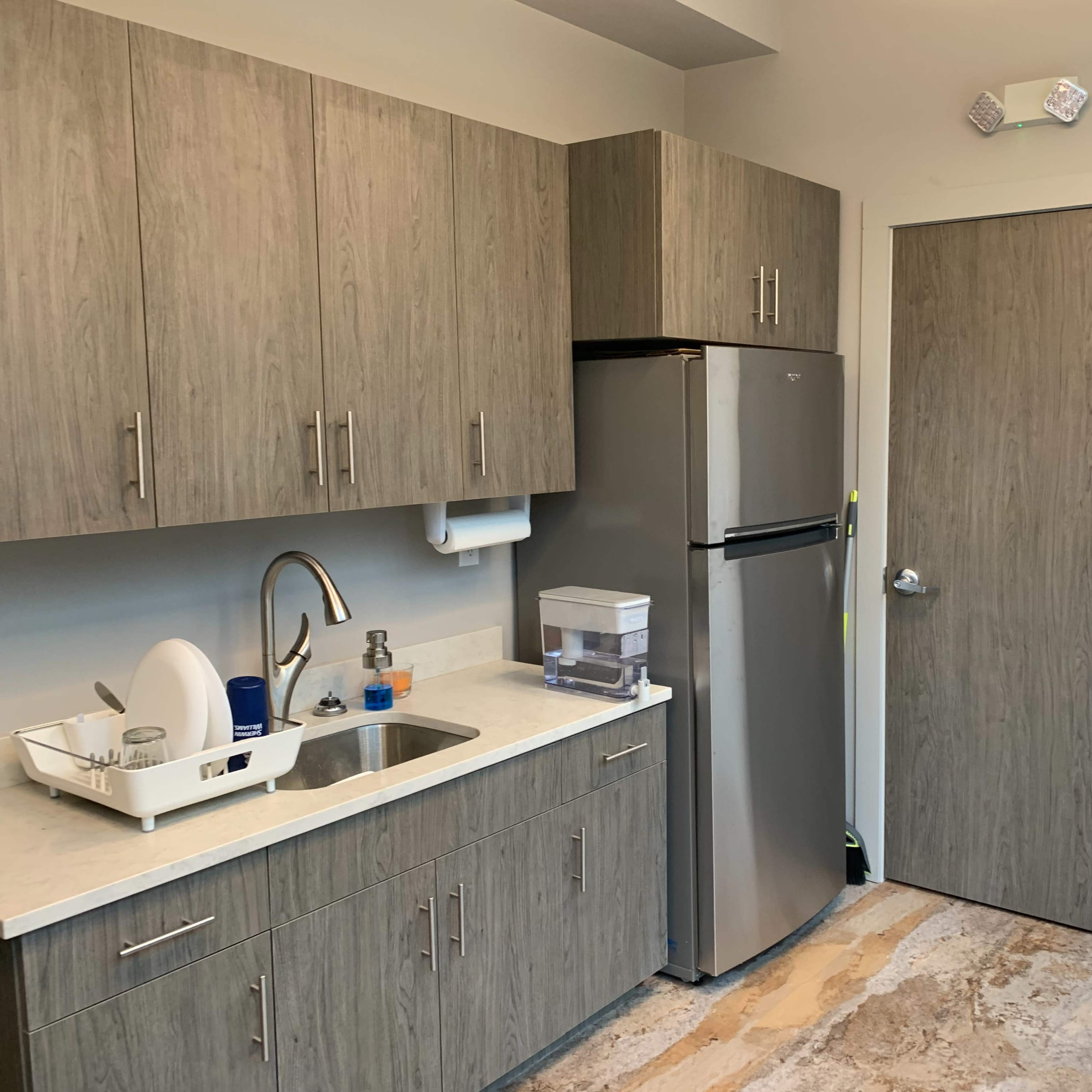 A modern kitchen area featuring gray cabinets, a stainless steel refrigerator, a sink, and a door leading to another room.