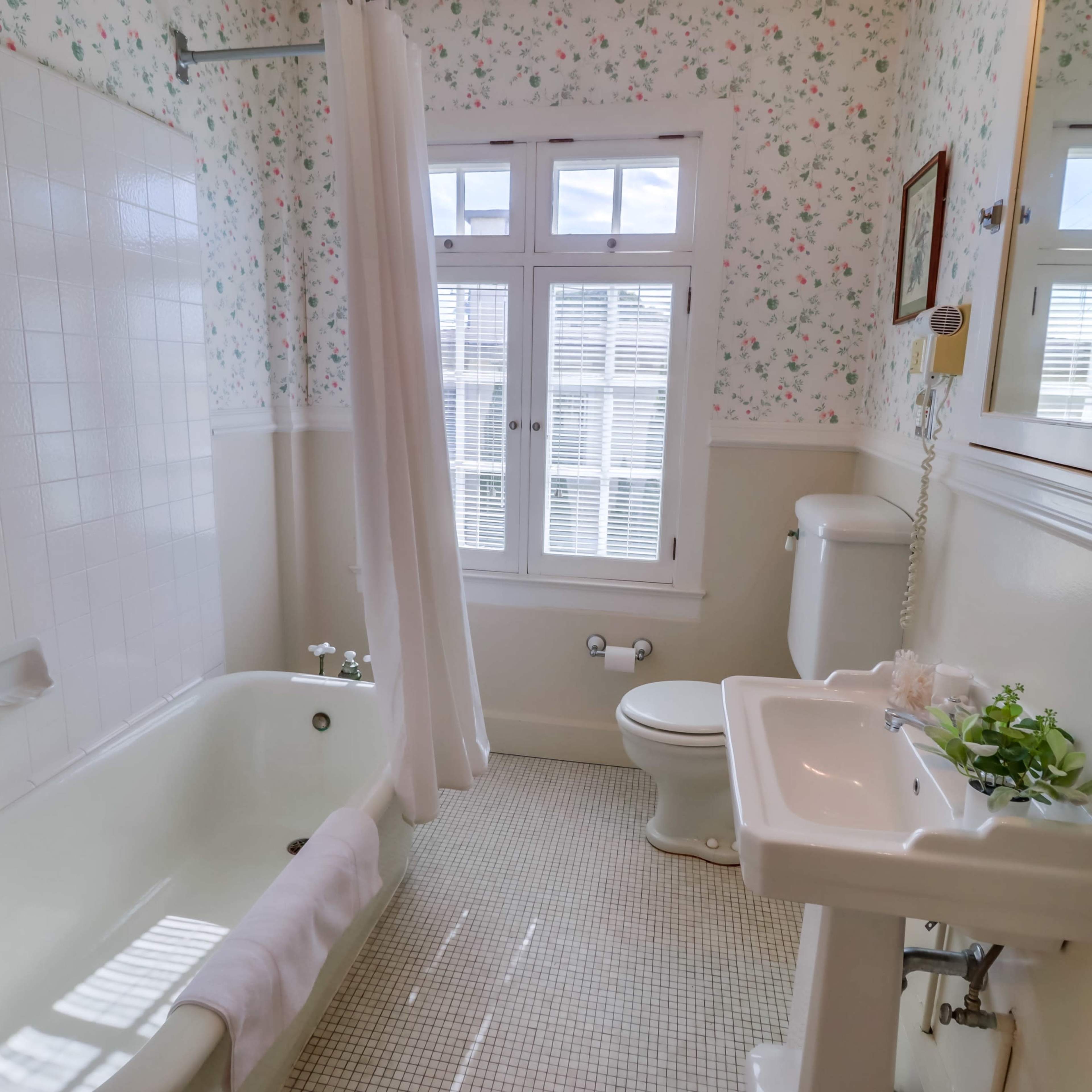 A bathroom with a bathtub, toilet, and sink, featuring floral wallpaper and a large window.