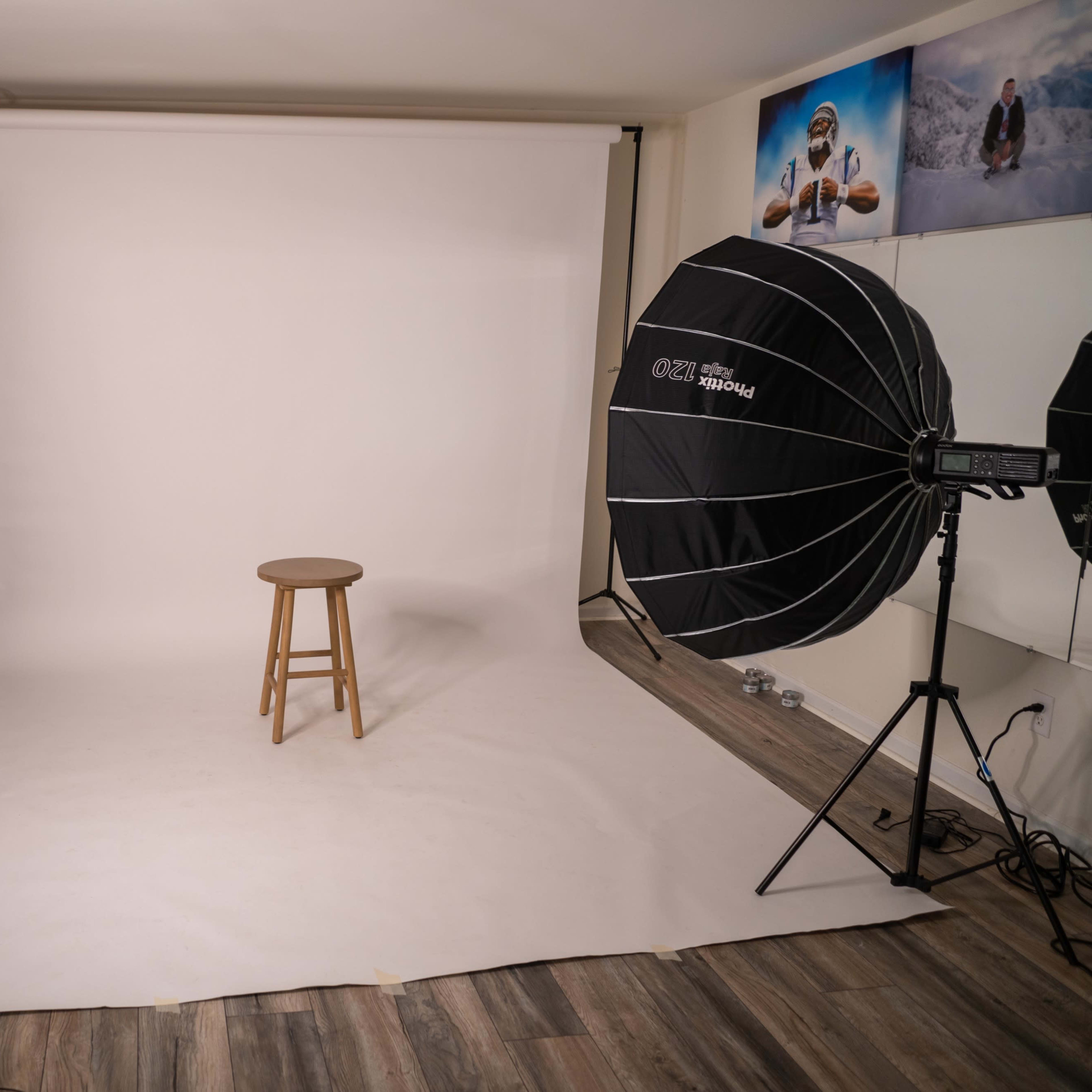 A photography studio setup with a blank backdrop, a wooden stool in the center, and various lighting equipment arranged around the perimeter.