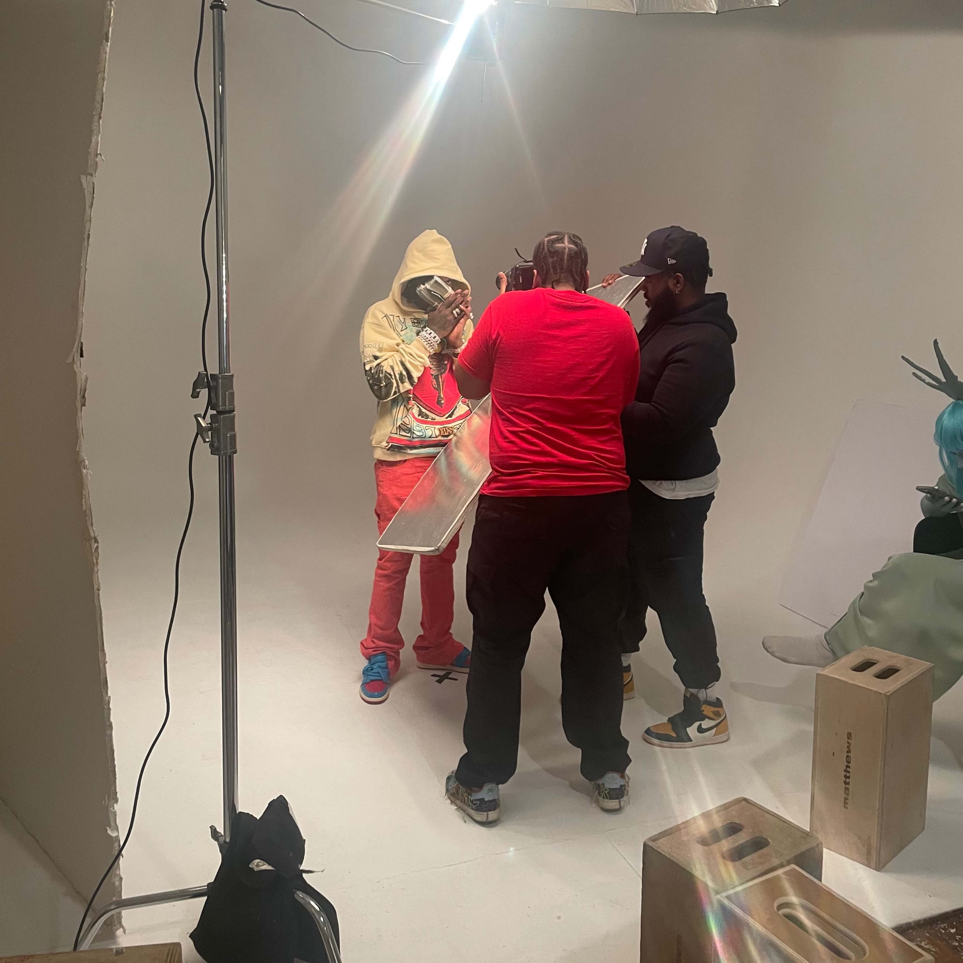 A group of three people collaborates in a photography studio, with one person dressed in a yellow hoodie being assisted with a prop under a large light source.
