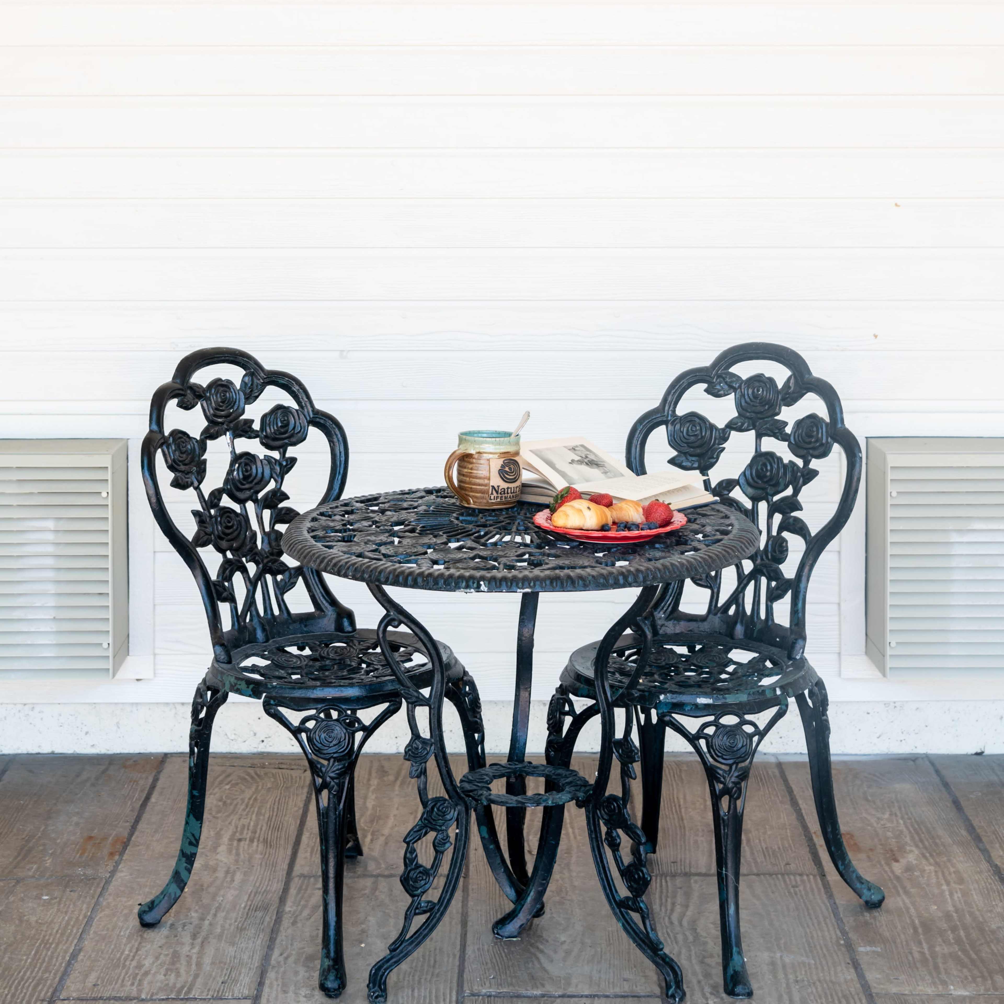 A wrought iron table with two matching chairs is set against a white wall, featuring a plate of apples and a mug on the table.