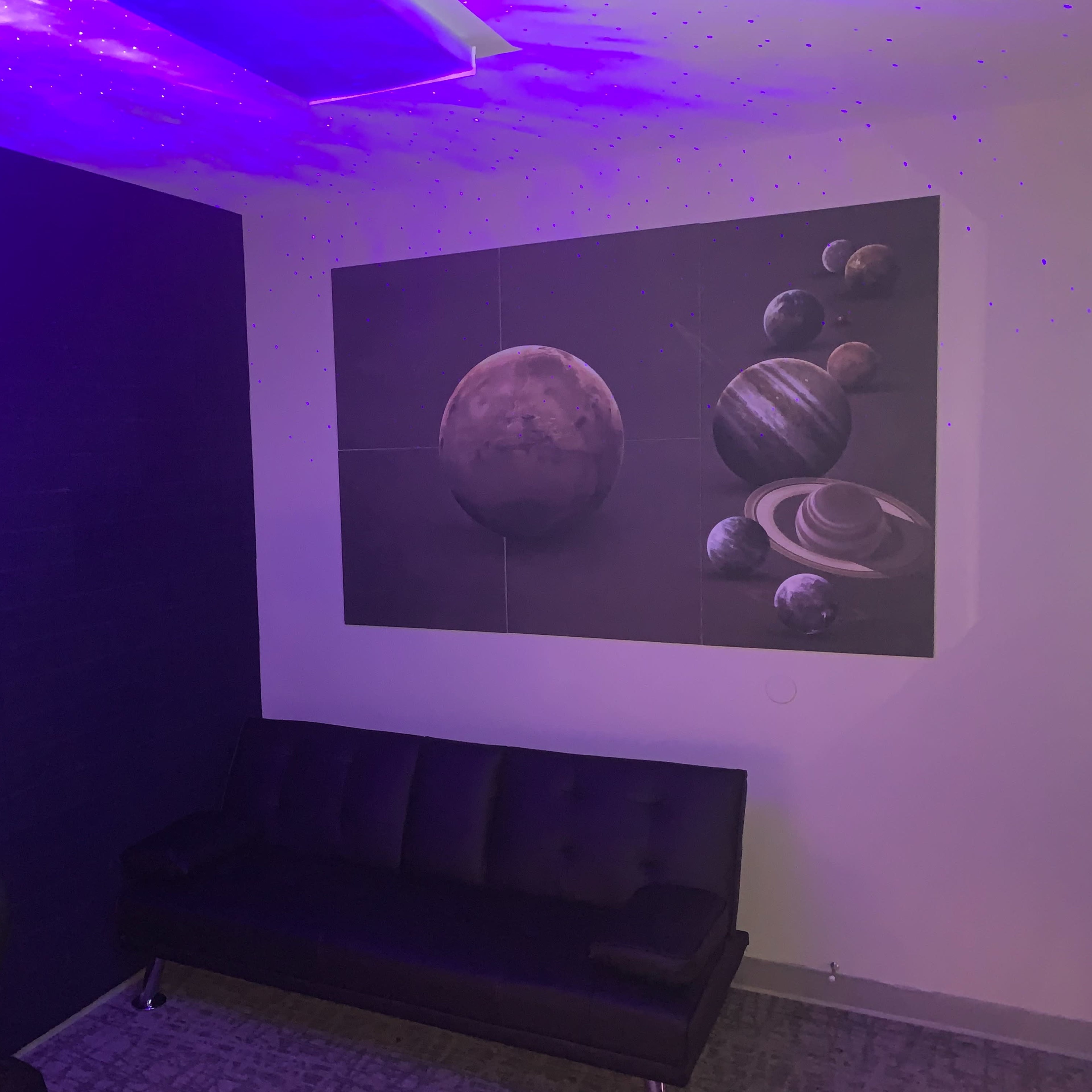 A room with a dark wall, a black couch, and a large mural of planets on the opposite wall, illuminated by purple lighting.