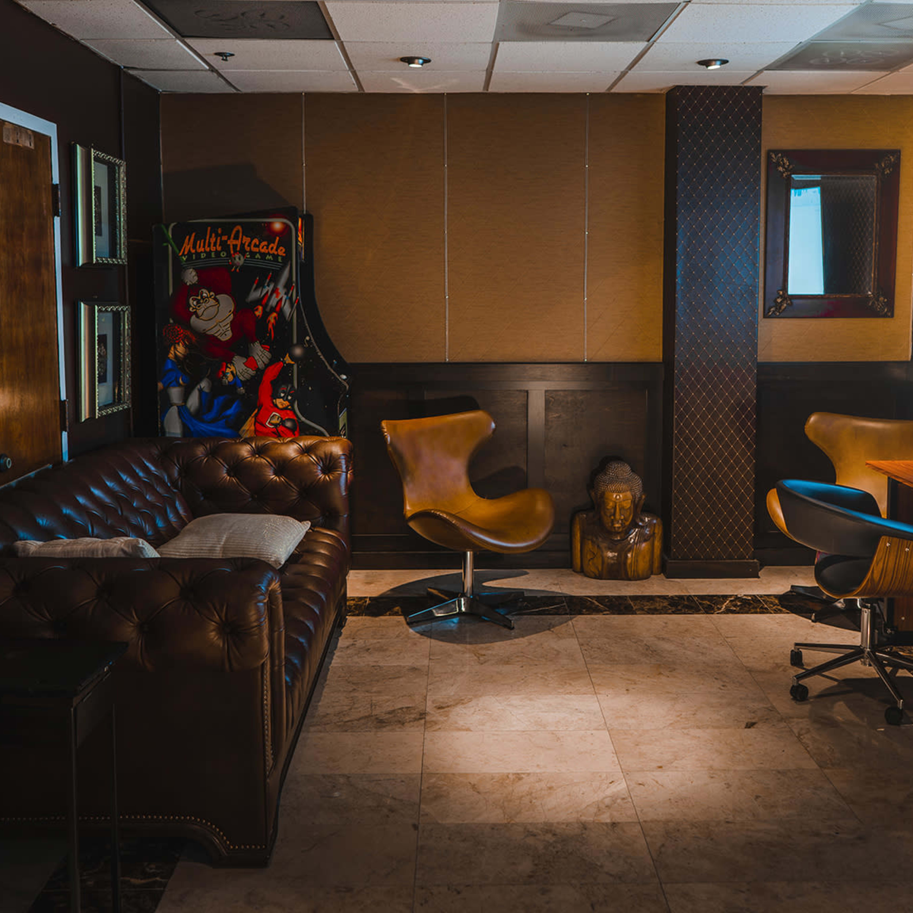 A small lounge area featuring a brown leather couch, two modern chairs, a wooden table with speakers, and a wall-mounted screen, all set against a backdrop of textured paneling.