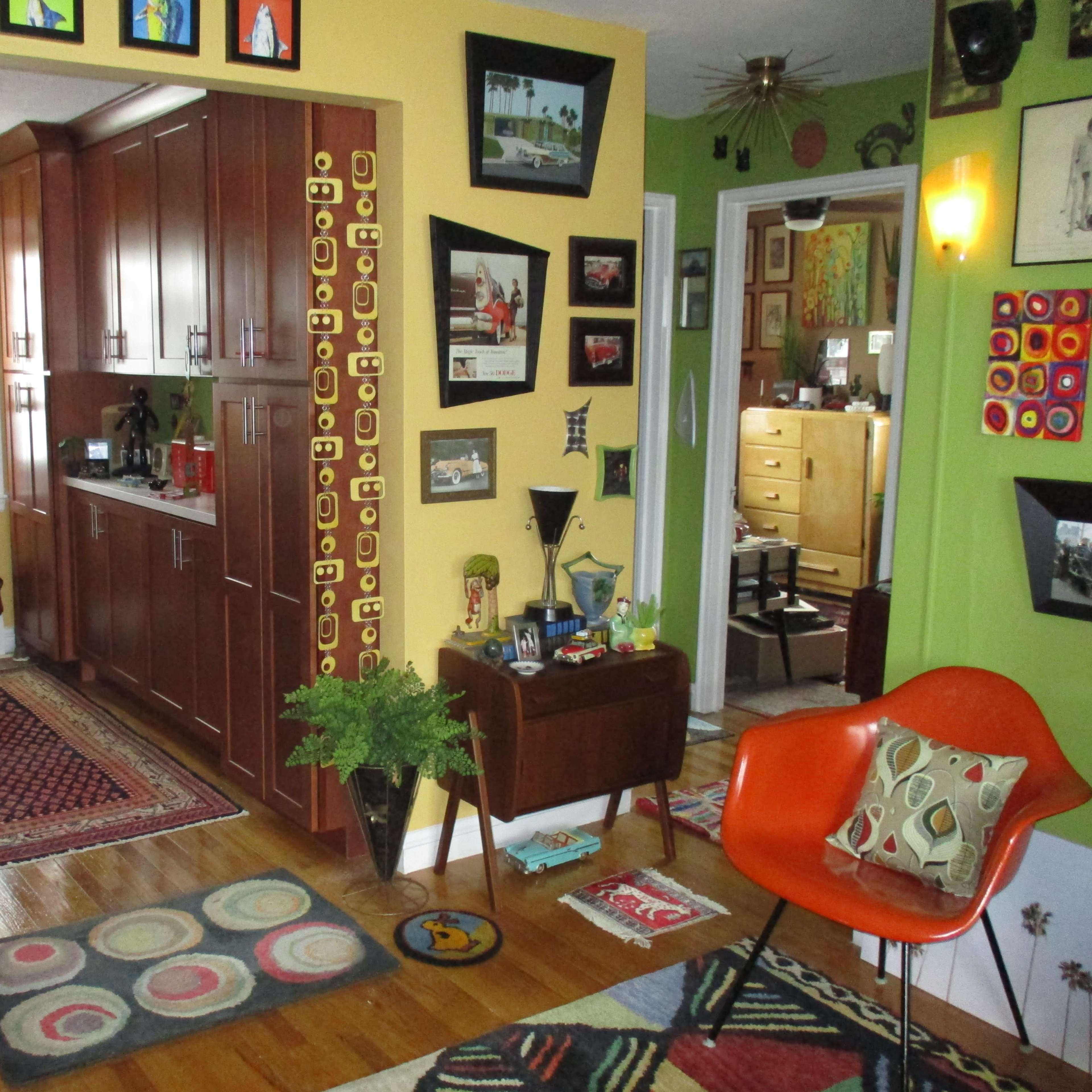 A brightly colored living space featuring a kitchen area, various framed pictures on the walls, a mid-century modern chair, and decorative rugs on wooden flooring.
