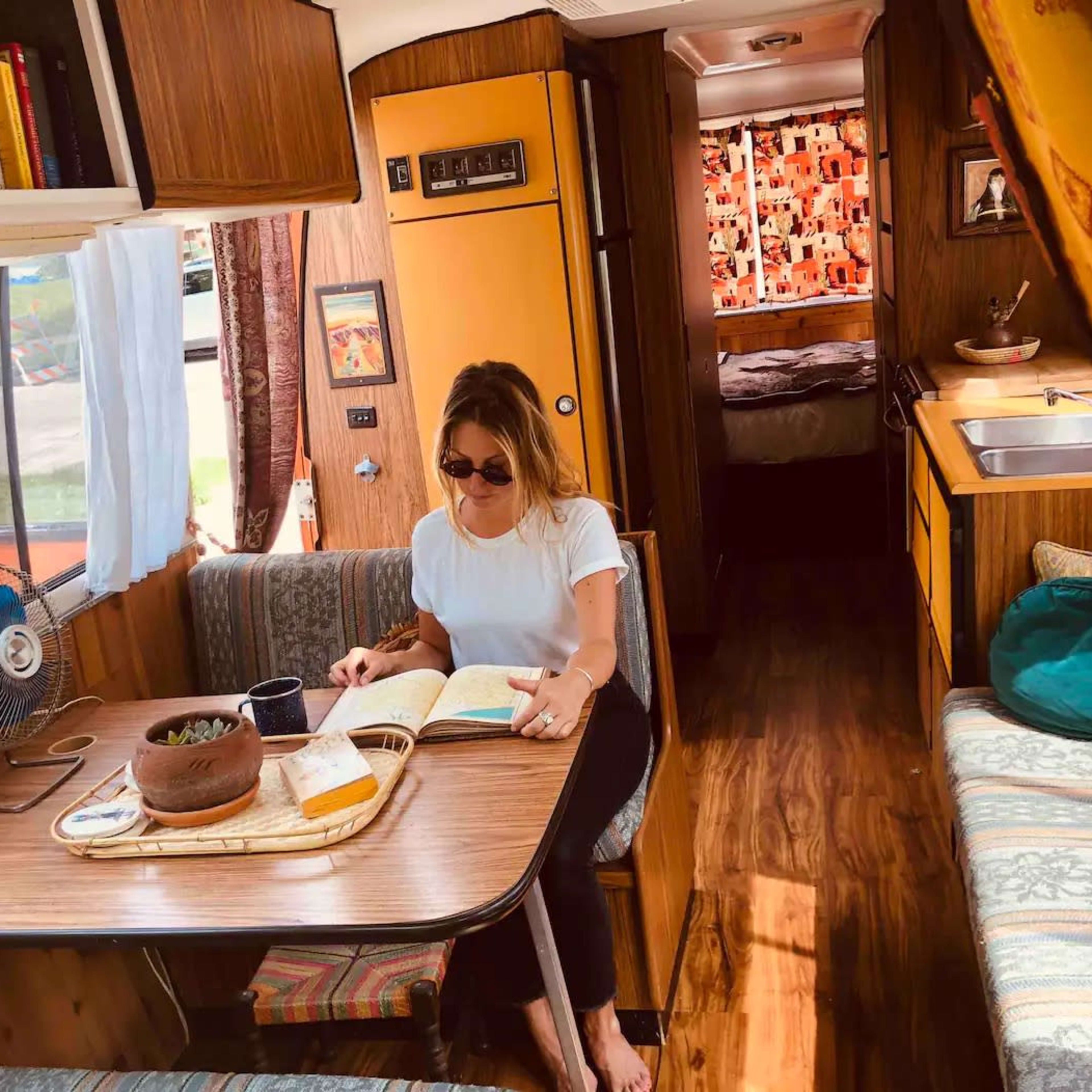 A woman sits at a table reading a book inside a vintage camper, surrounded by wooden paneling and colorful cushions.