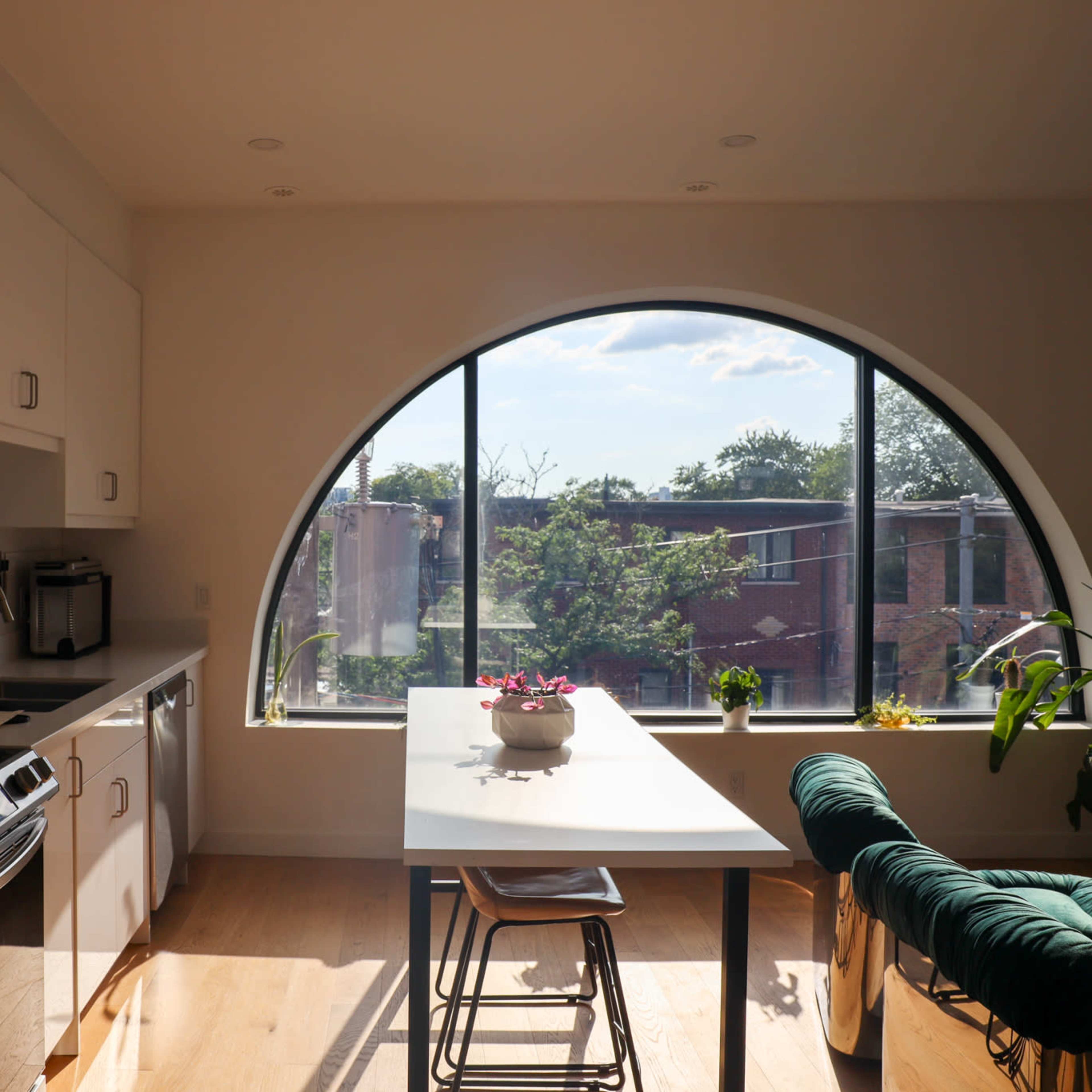 A modern kitchen features a bright, arched window overlooking a landscaped outdoor area, with a dining table and chairs positioned nearby.