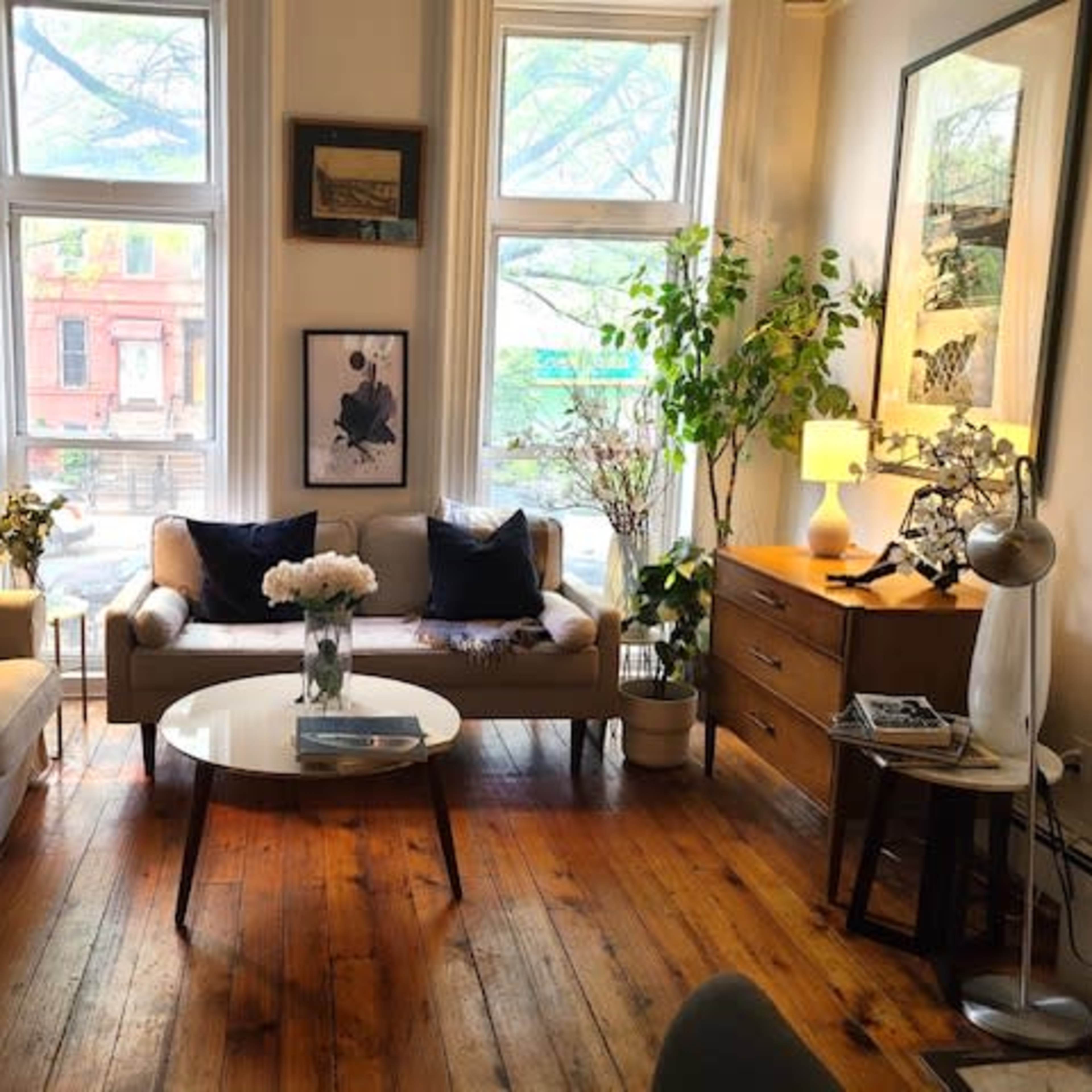 A cozy living room featuring two sofas, a round coffee table, and a wooden side table, with large windows letting in natural light and plants placed throughout the space.