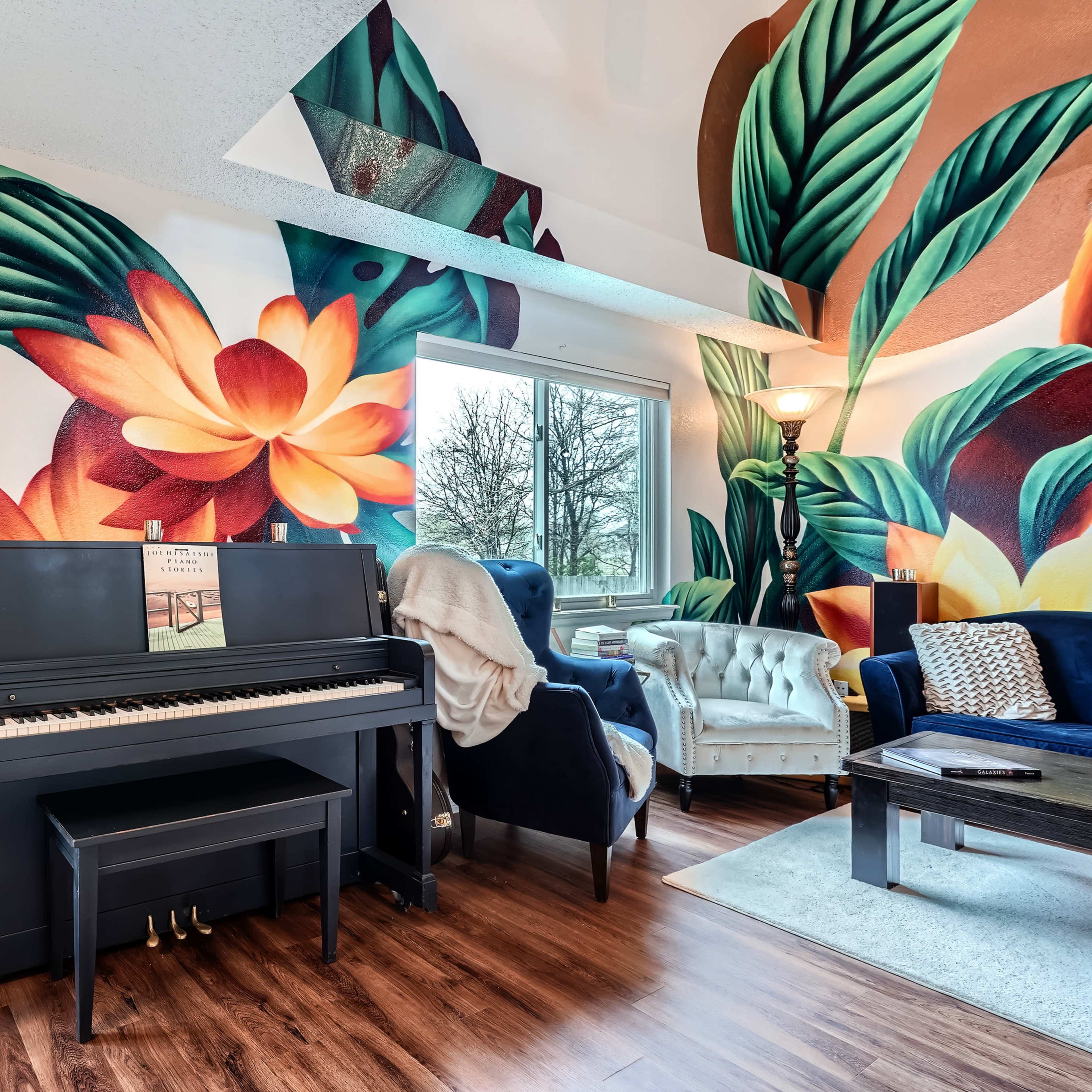 A black piano is positioned against a wall adorned with vibrant tropical-themed murals, featuring large leaves and colorful flowers, in a room with a large window and stylish furniture.