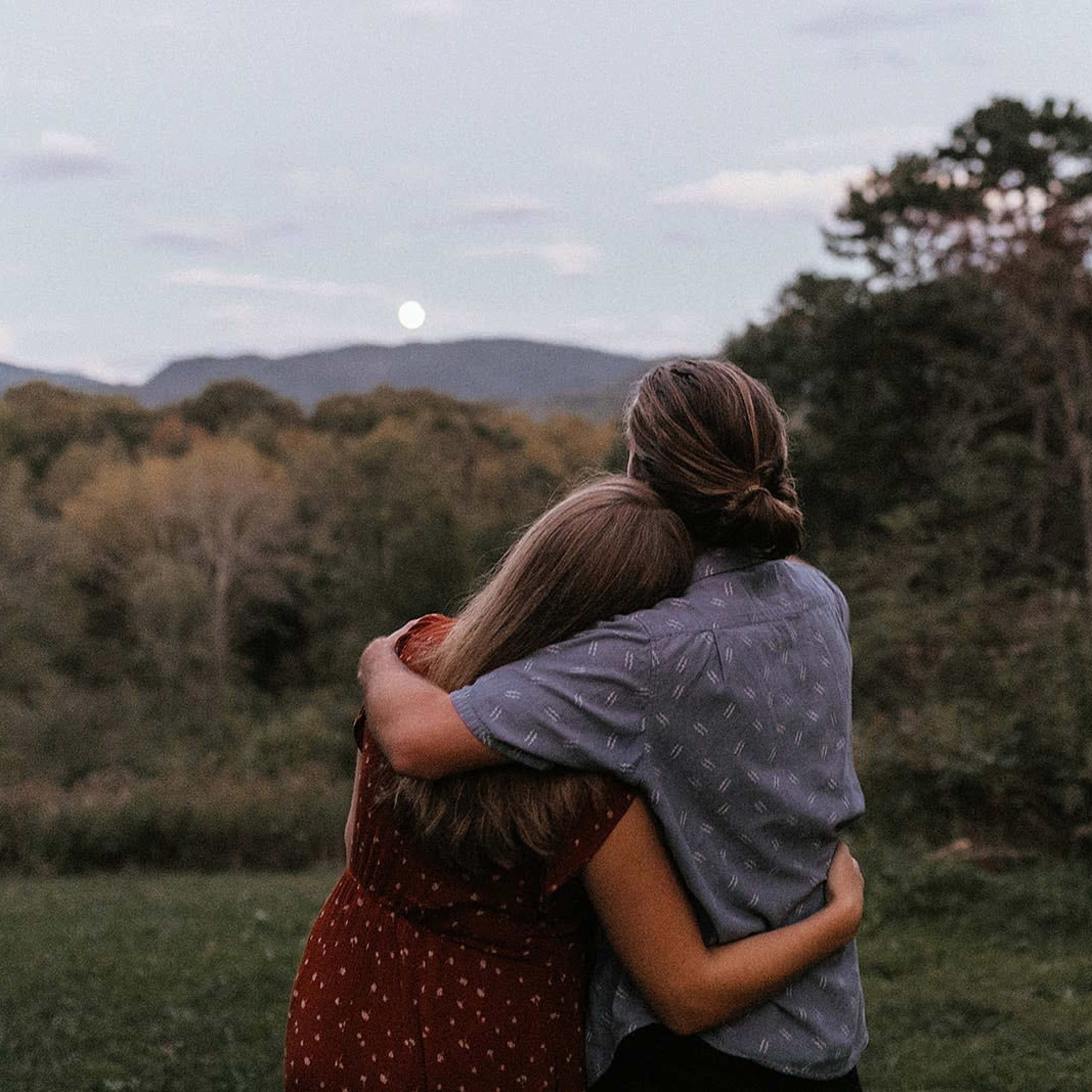 A couple stands together in an open field, embracing while facing a mountain landscape at dusk.