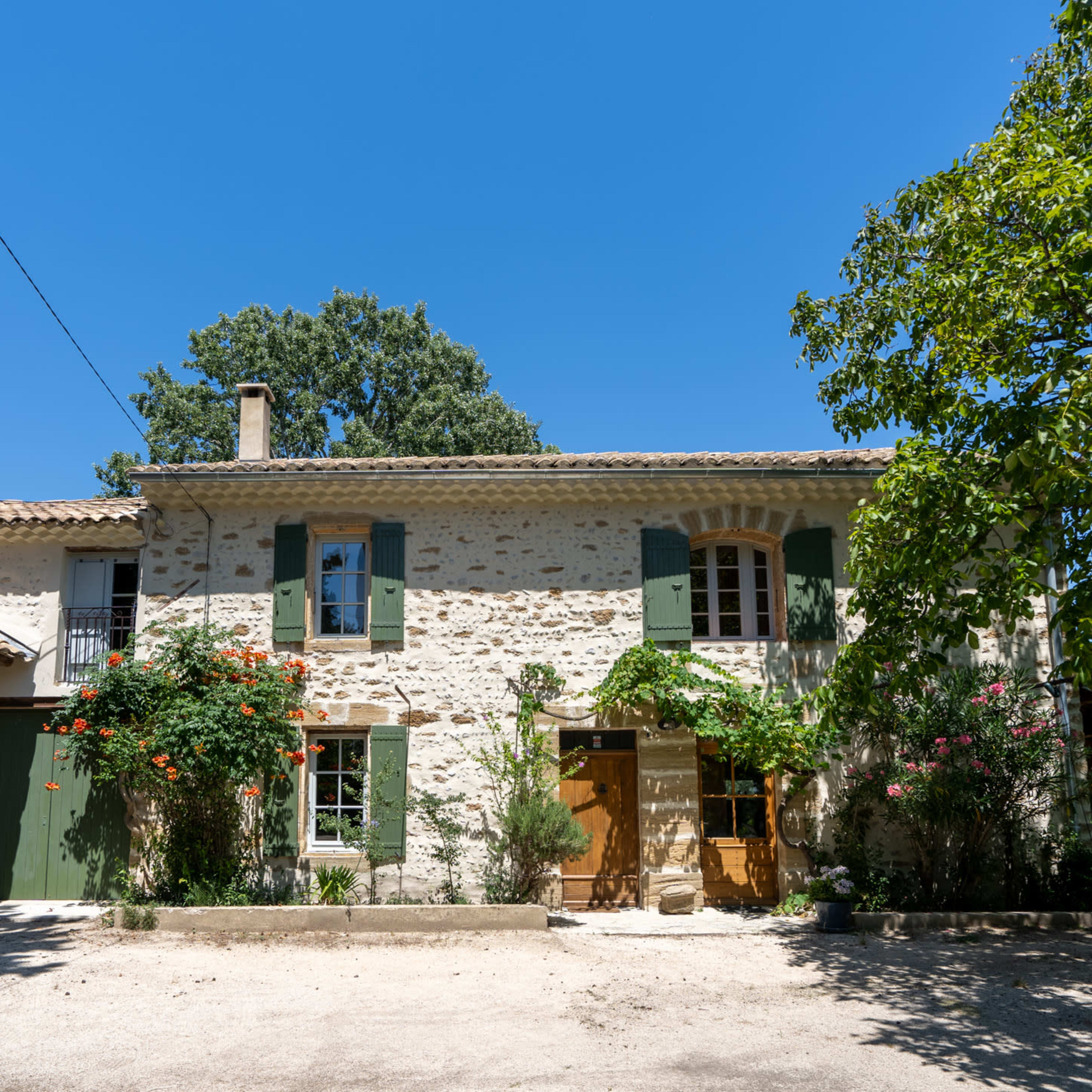 A stone house with green shutters and a wooden door is surrounded by trees and flowering plants under a clear blue sky.