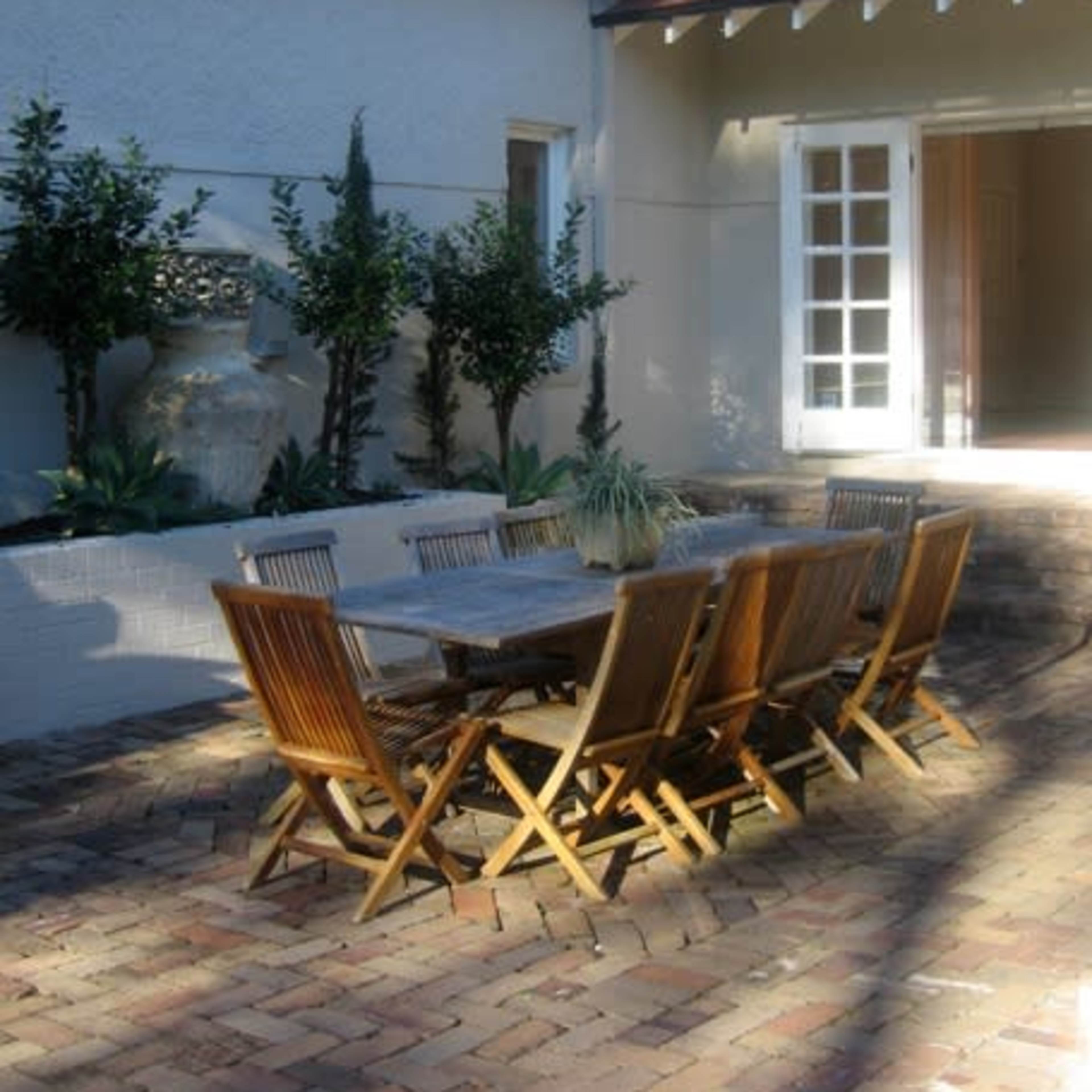 A wooden dining table with chairs is set up in a brick-paved courtyard beside a house.