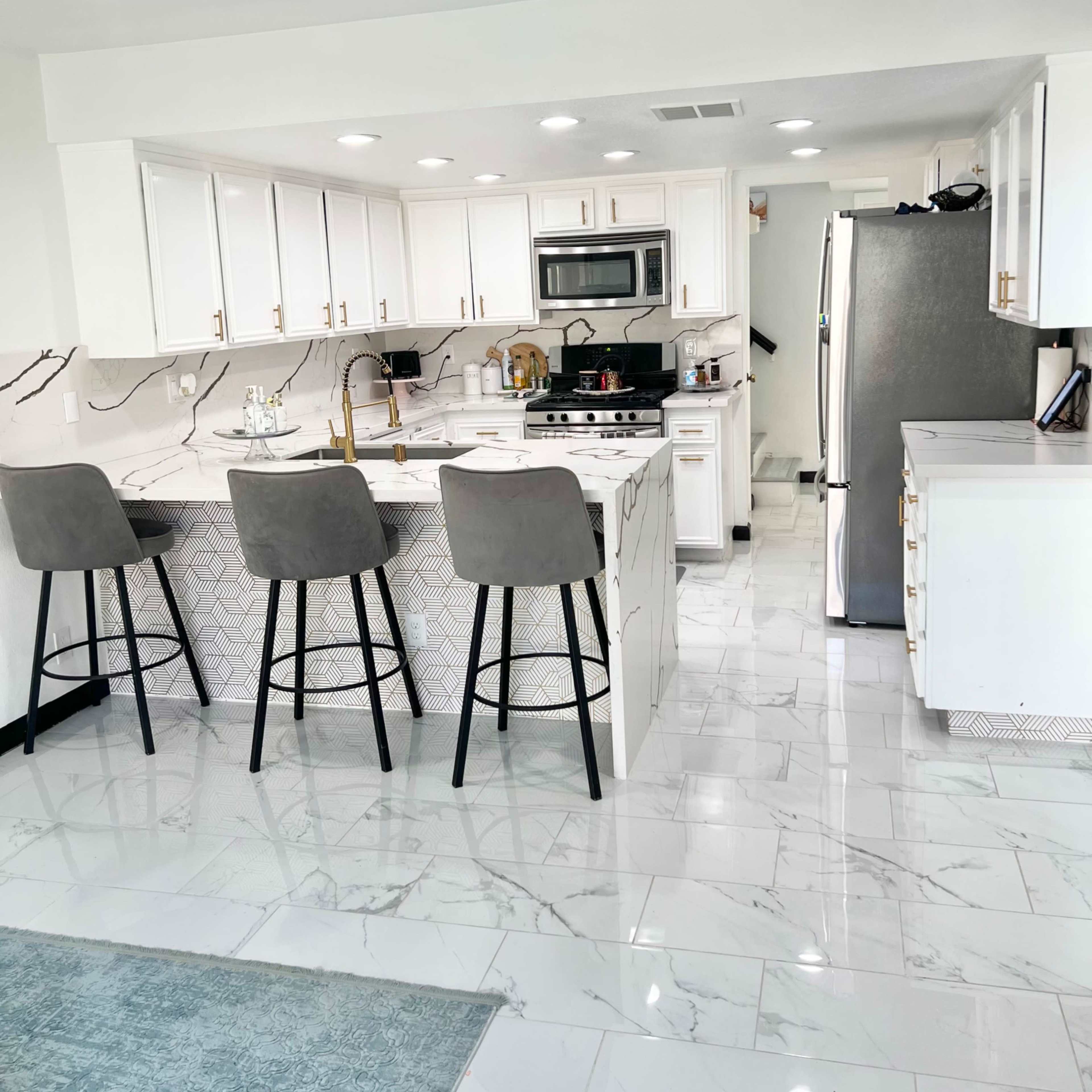 A modern kitchen with white cabinets, gray bar stools, stainless steel appliances, and a marble-patterned backsplash.