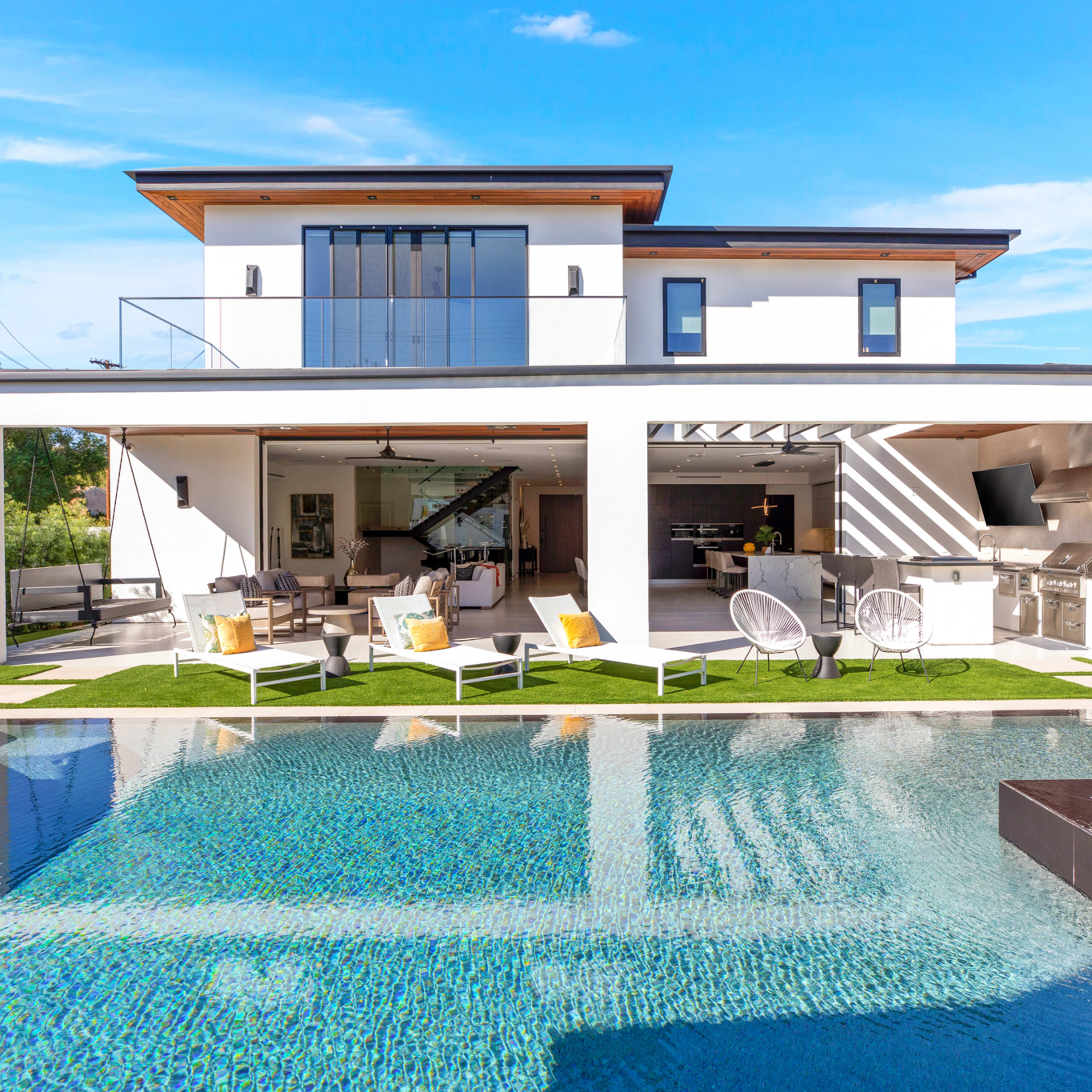 A modern two-story house features an outdoor patio with seating and a pool surrounded by landscaped greenery.
