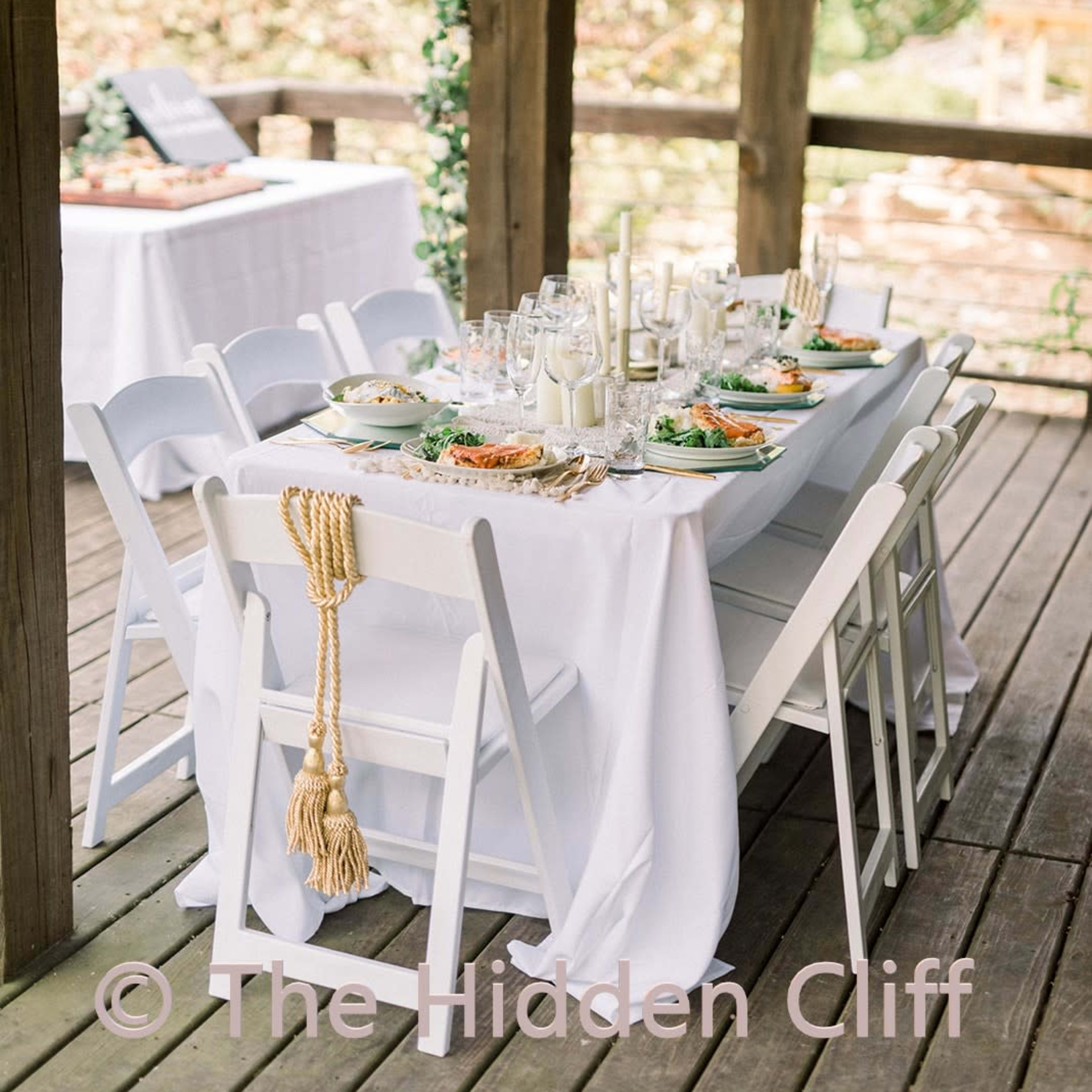 A long dining table is set with plates of food and drinks under a wooden gazebo, surrounded by strings of lights and greenery.