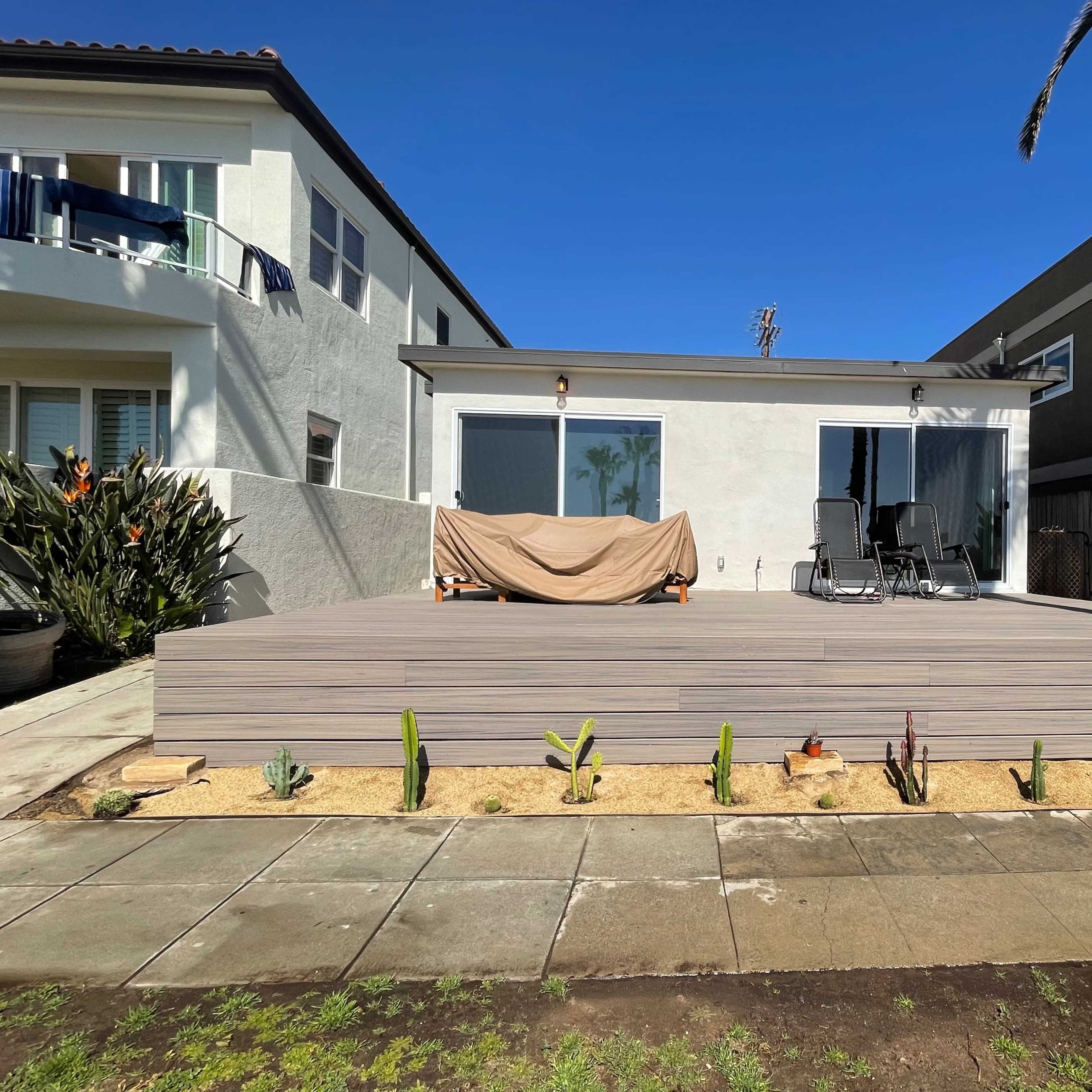 A large wooden deck in front of a house, featuring two chairs and a covered area, with a landscaped garden of cacti in the foreground.
