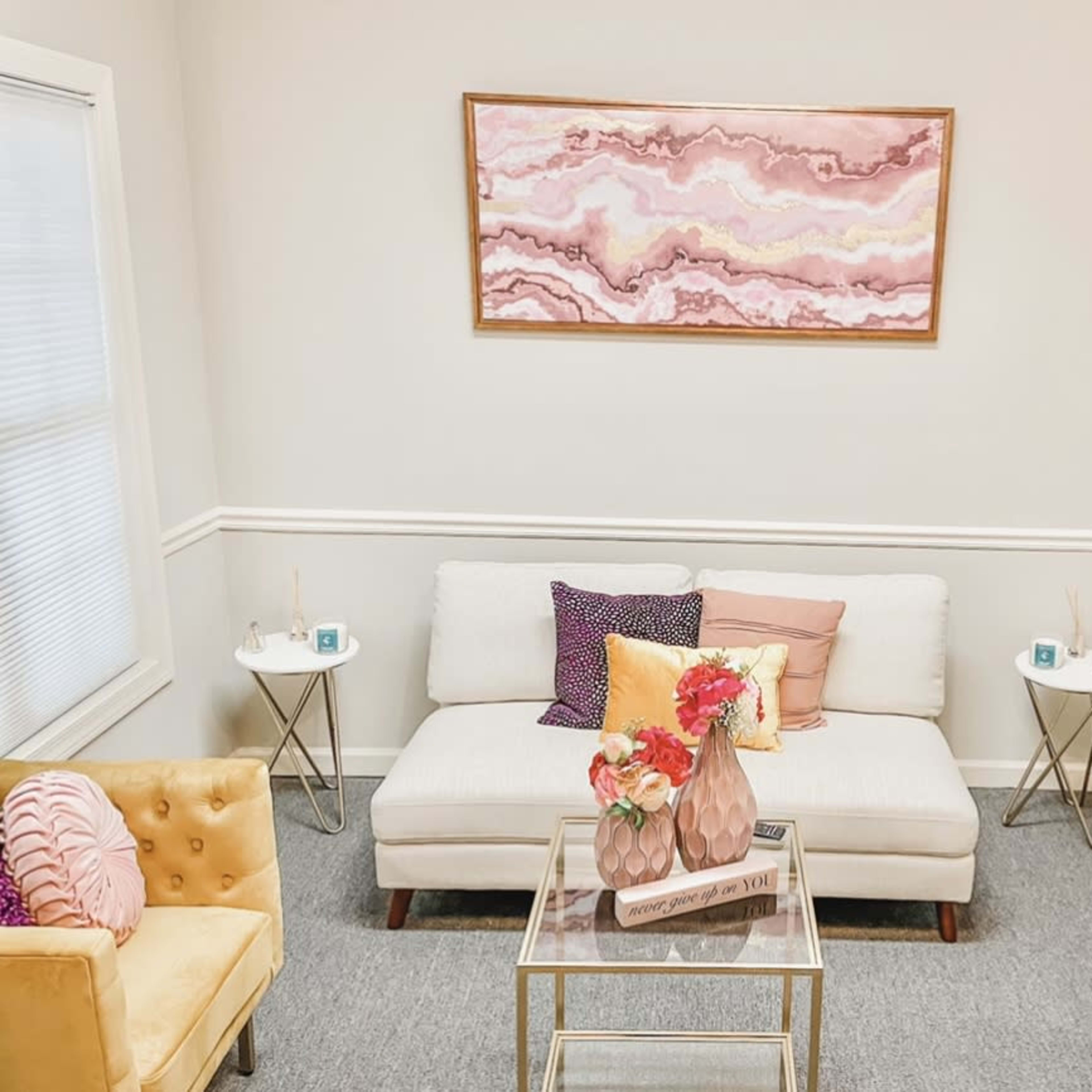 A small, elegantly arranged waiting area with a white sofa, two yellow chairs, a glass coffee table, and a large wall art piece above.