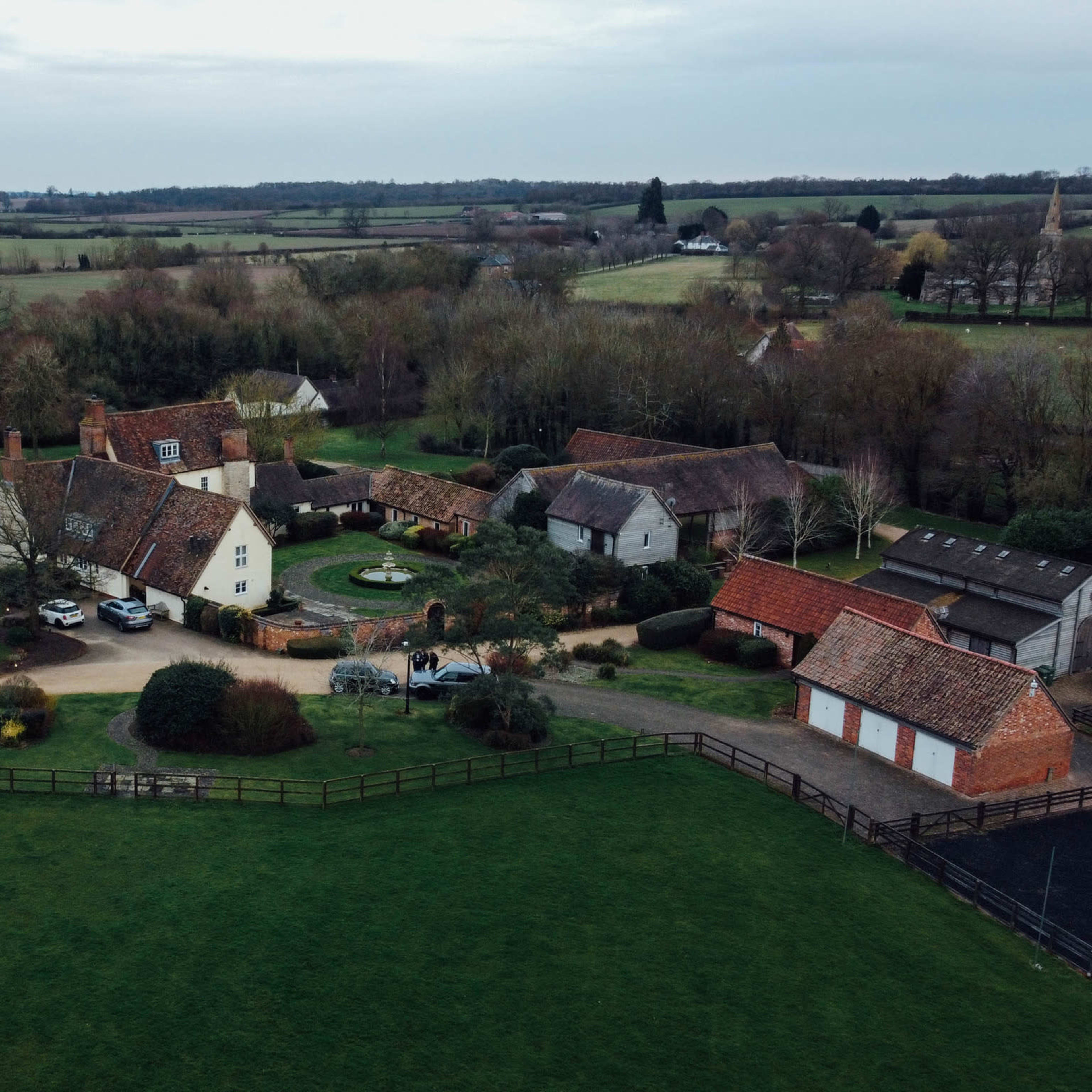 An aerial view shows a cluster of farmhouses and outbuildings surrounded by green fields and trees.