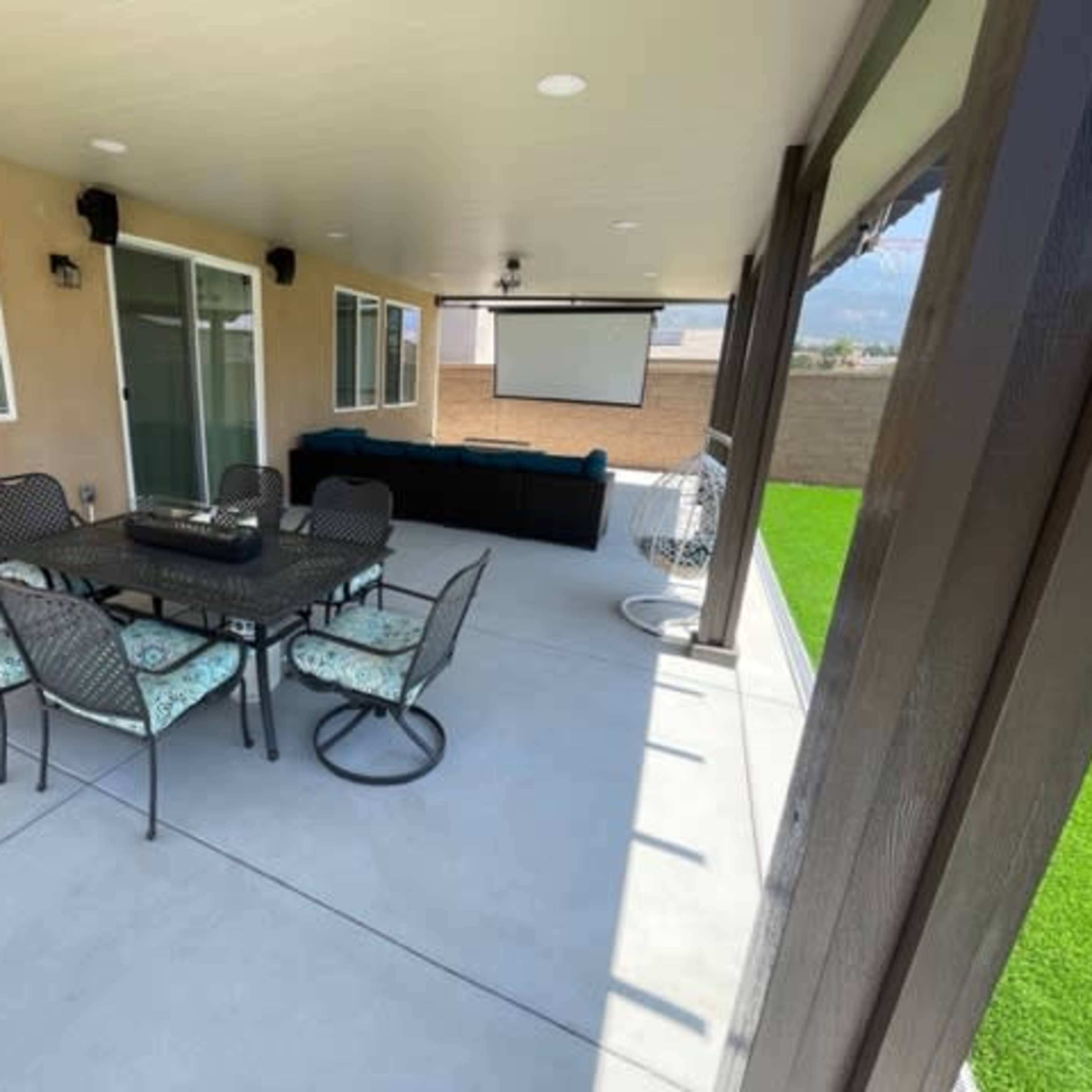 A covered outdoor patio with a dining table and chairs, a couch, and a projector screen mounted on the wall.