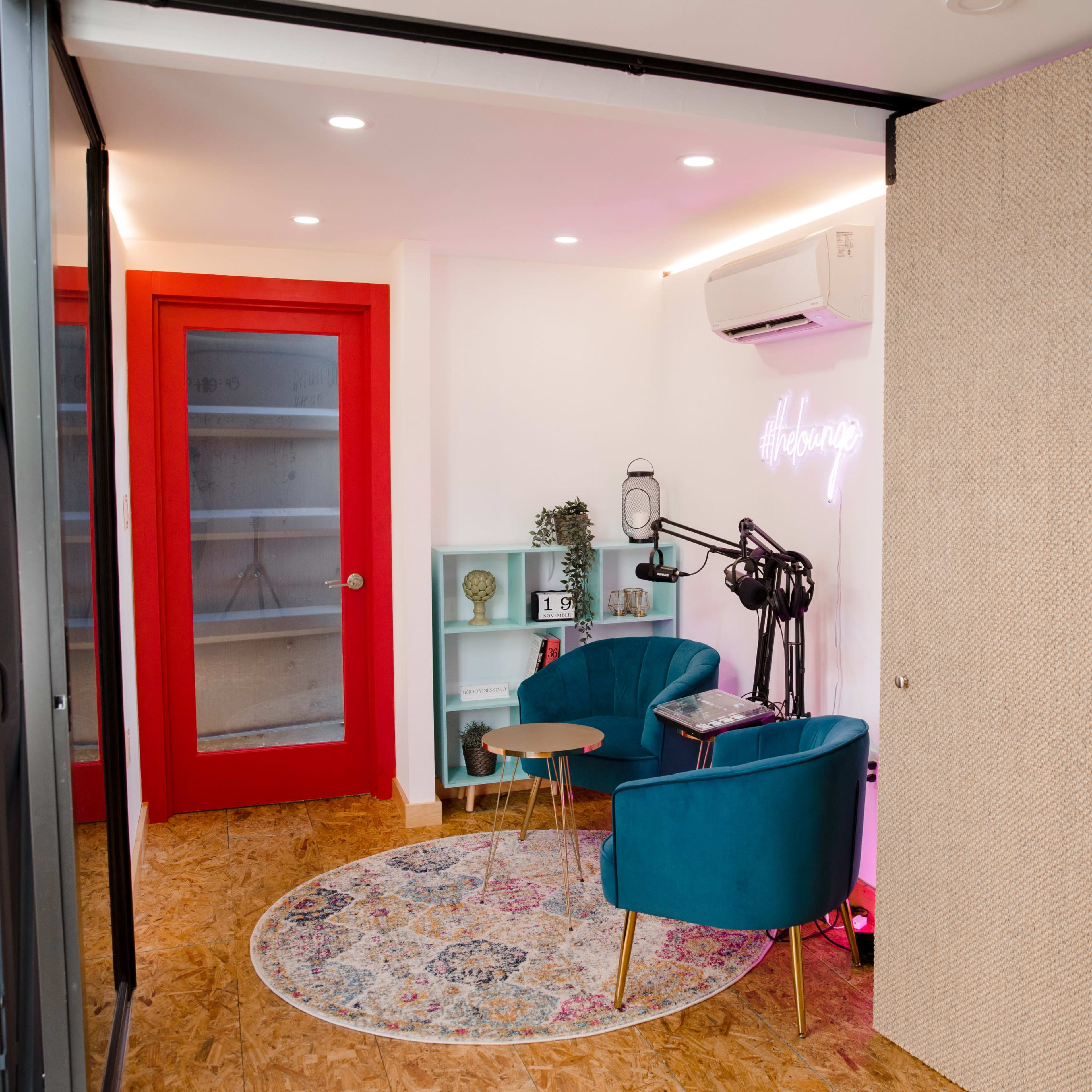 A modern room with two teal chairs, a small wooden table, and a colorful rug, featuring a red glass door and decorative shelves.