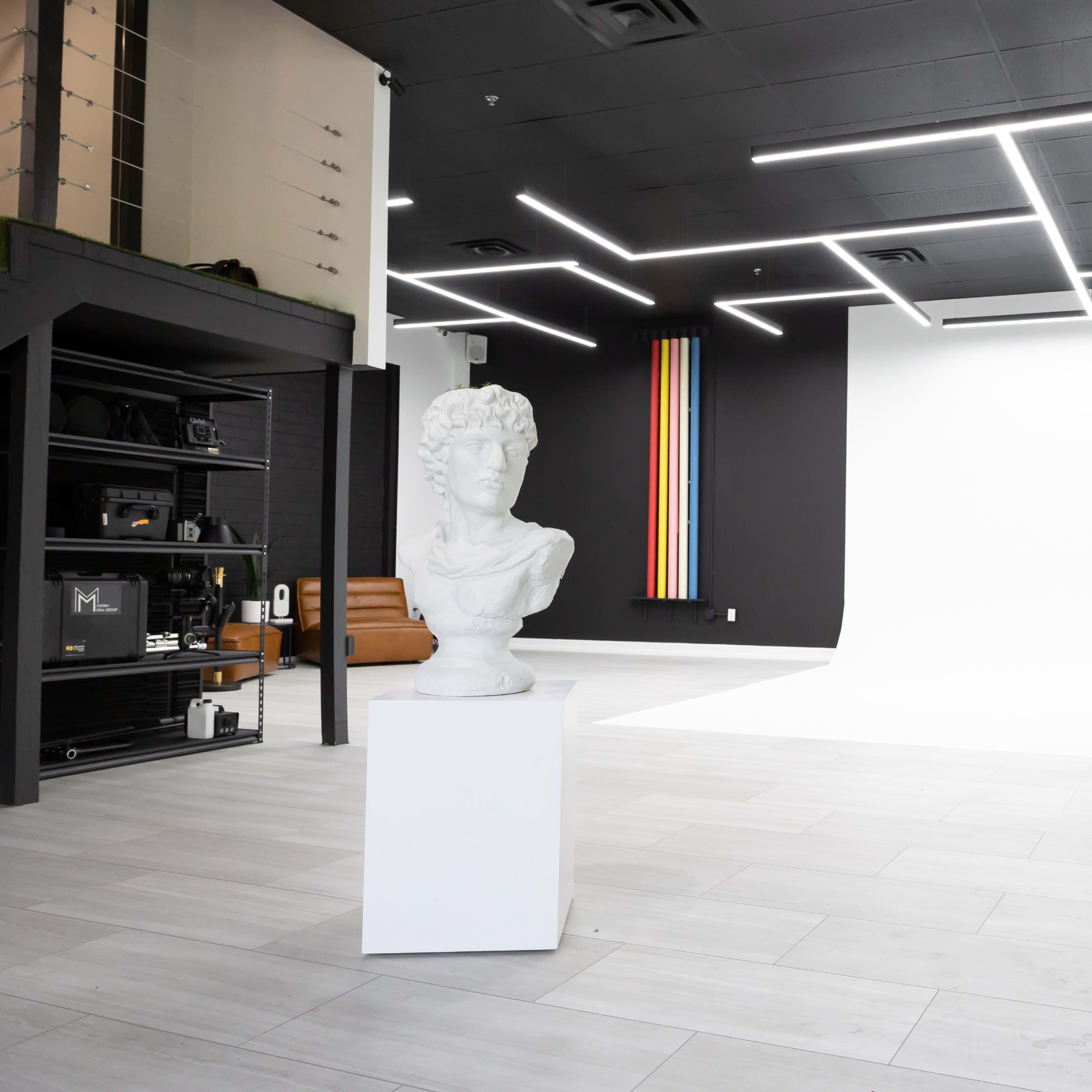 A modern studio space featuring a white backdrop, a contemporary sculpture on a pedestal, and green plants in black pots.