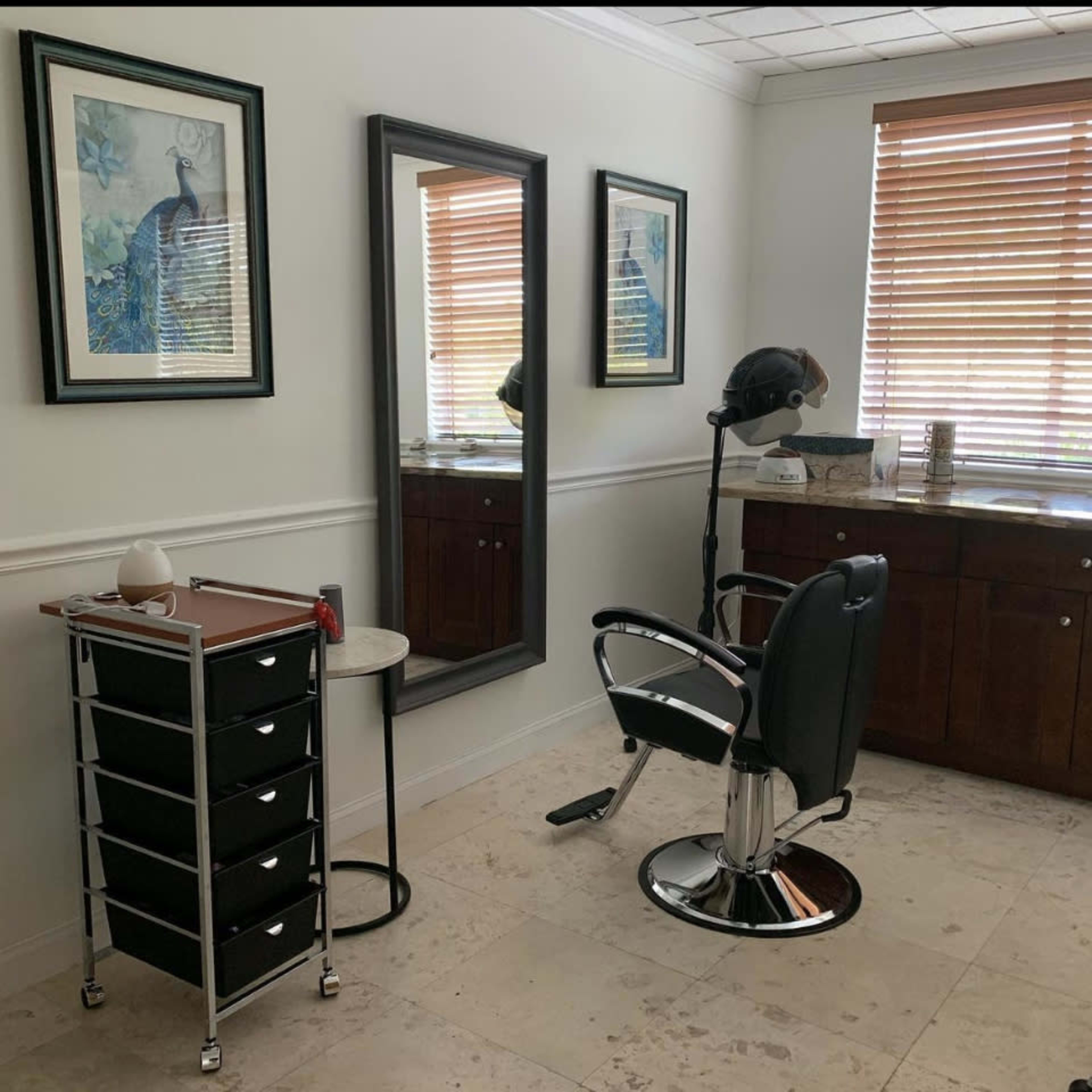 A hair salon room features a black salon chair, a mirror, a storage cart with drawers, and framed artwork on the walls.