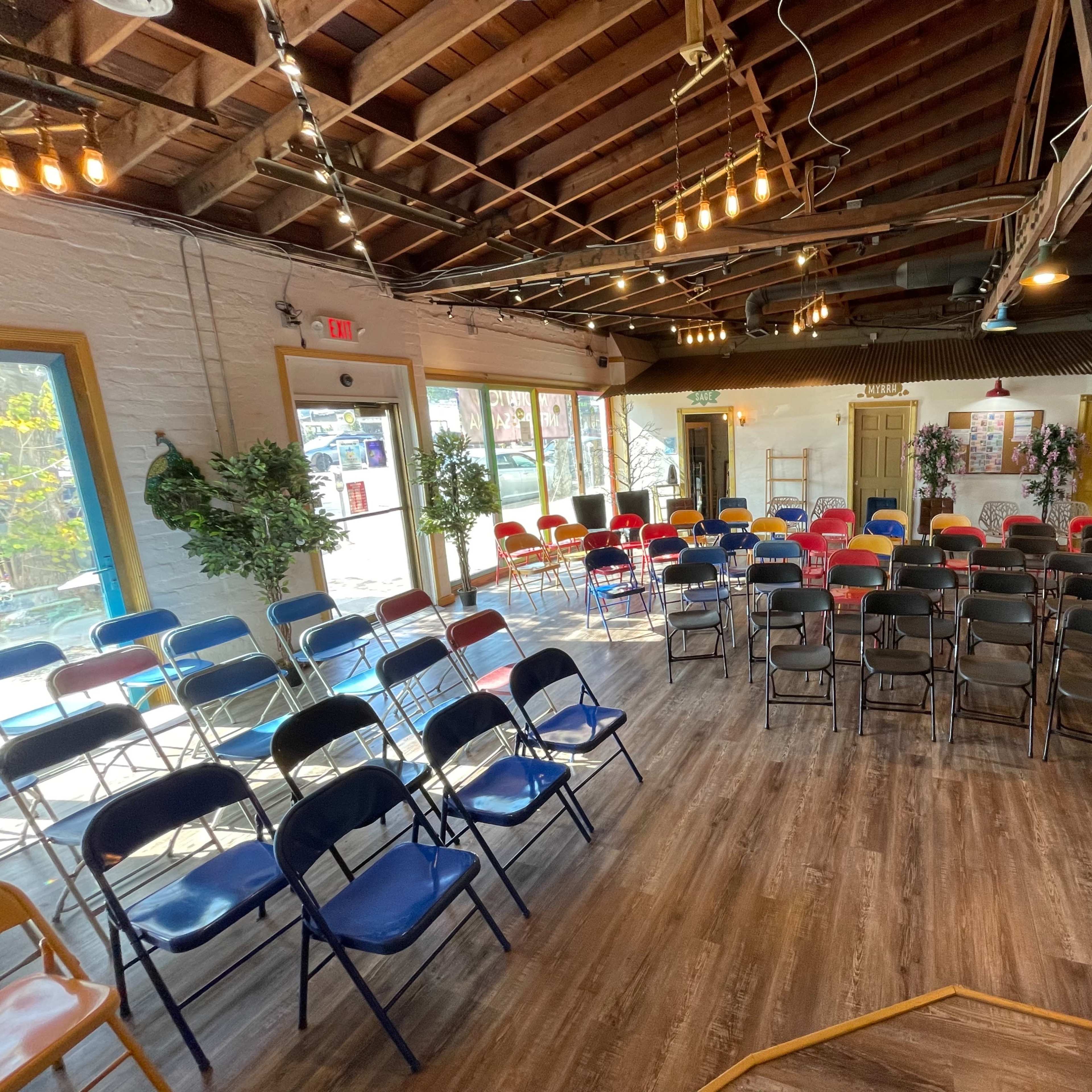 A spacious room with rows of multicolored folding chairs arranged for an event, under a wooden ceiling with exposed beams and hanging lights.