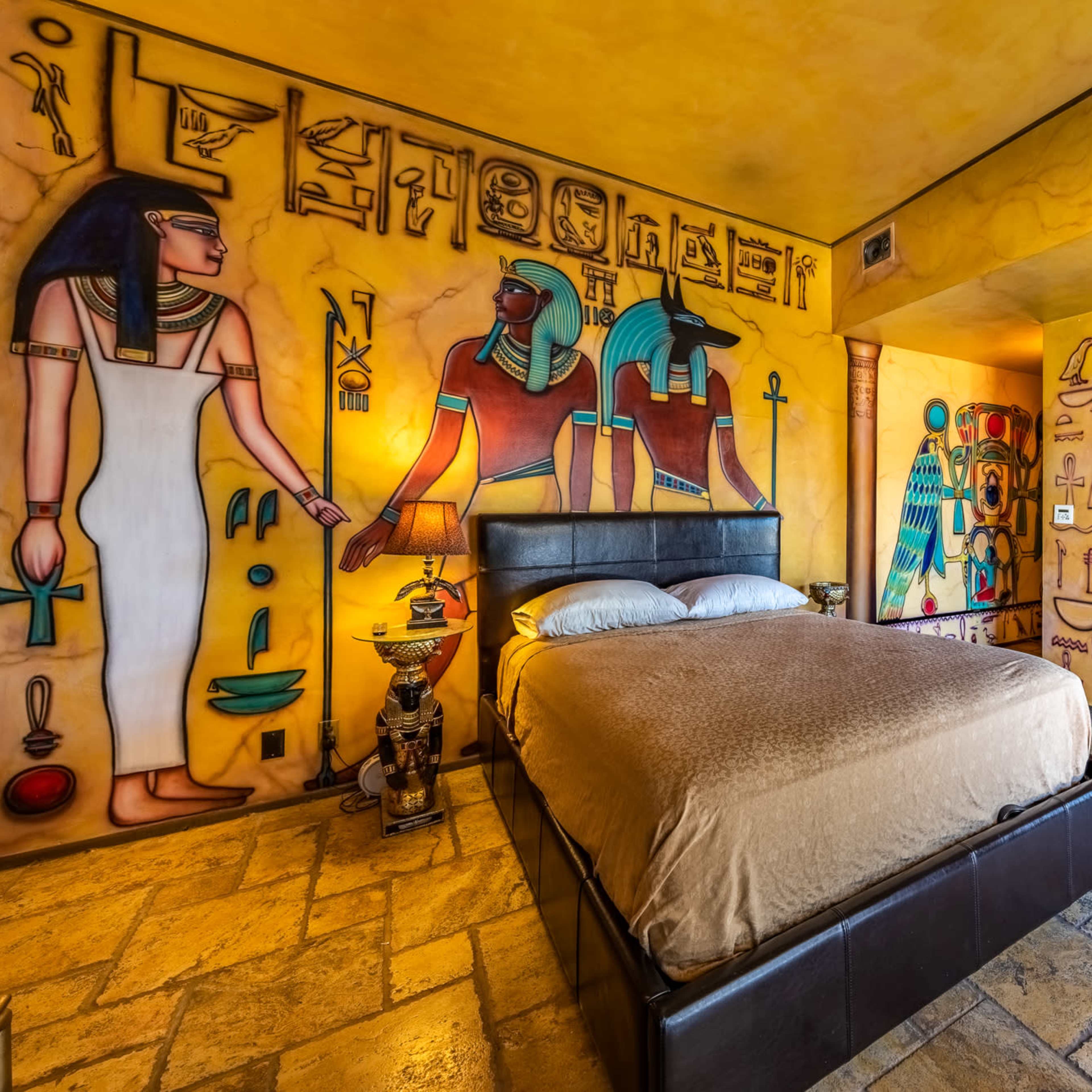 The scene depicts a bedroom with walls decorated in an ancient Egyptian theme, featuring hieroglyphs and images of Egyptian figures, along with a bed centrally placed in the room.