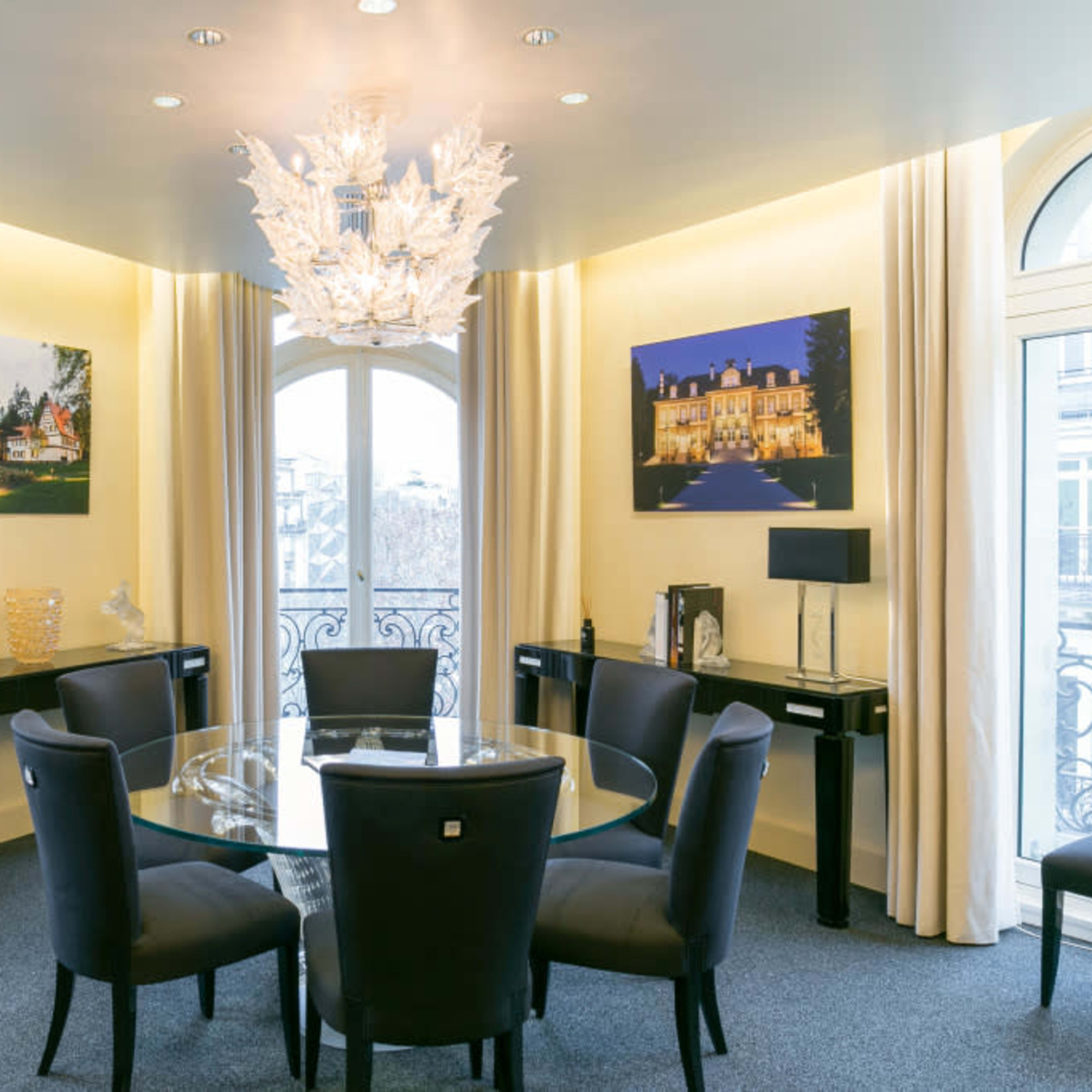 A modern meeting room with a glass table surrounded by black chairs, featuring large windows and two framed photographs on the walls.
