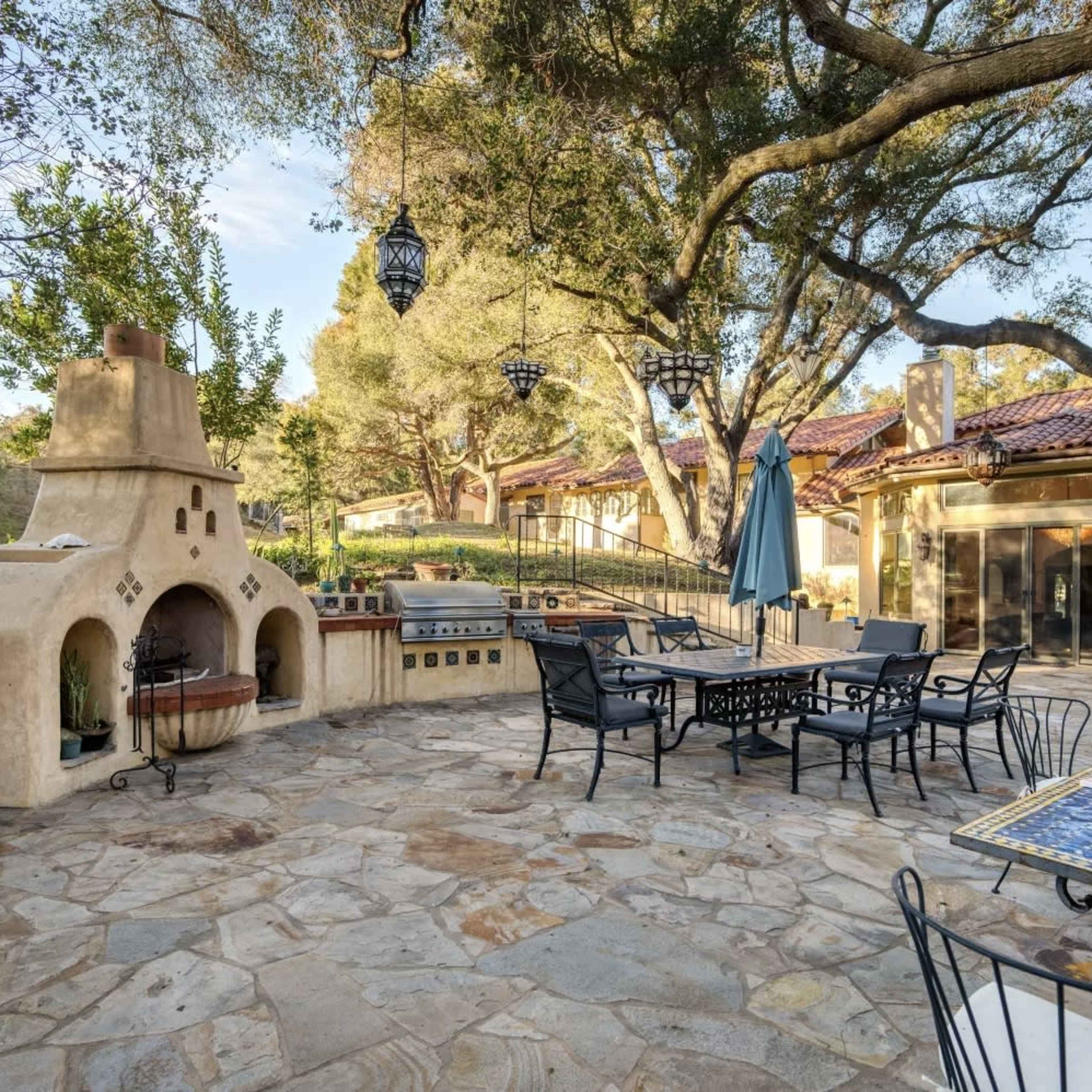 A spacious outdoor patio area featuring a large adobe-style fireplace, stone flooring, and black metal furniture surrounded by trees and decorative lanterns.