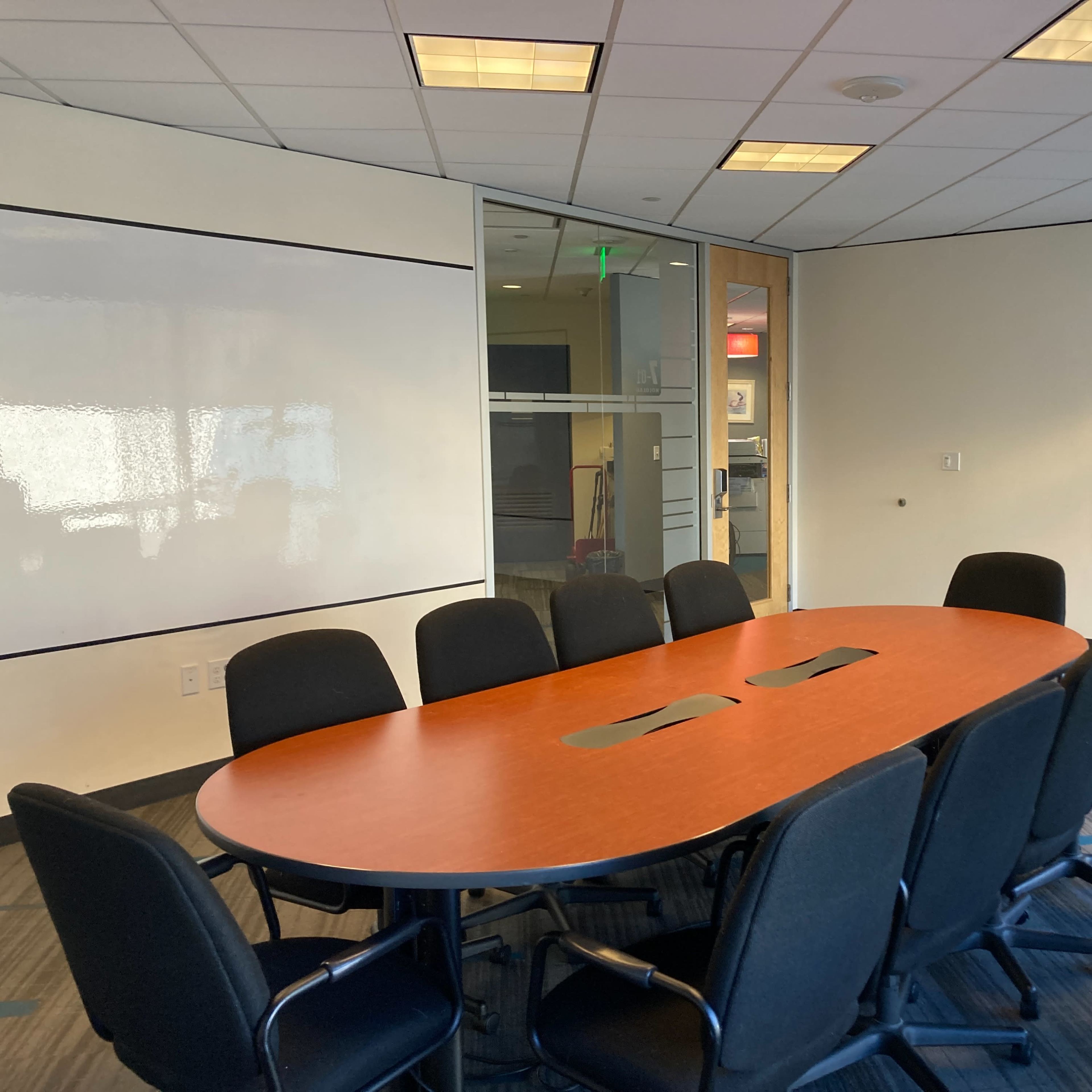 A conference room features a large oval table surrounded by black chairs, with whiteboards on two walls and a glass door leading to an adjoining space.