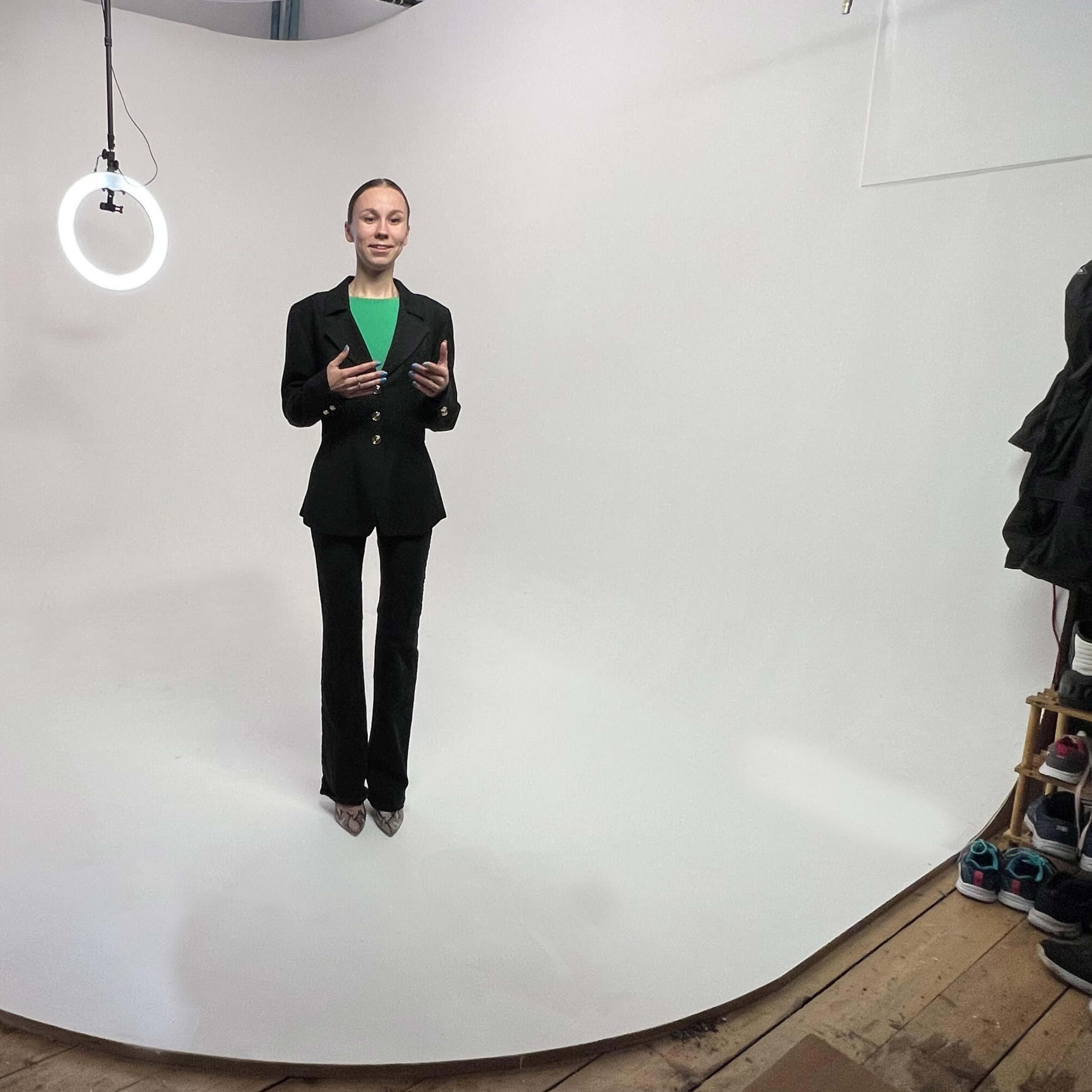 A woman in a black suit stands on a white backdrop, with a ring light suspended above her.