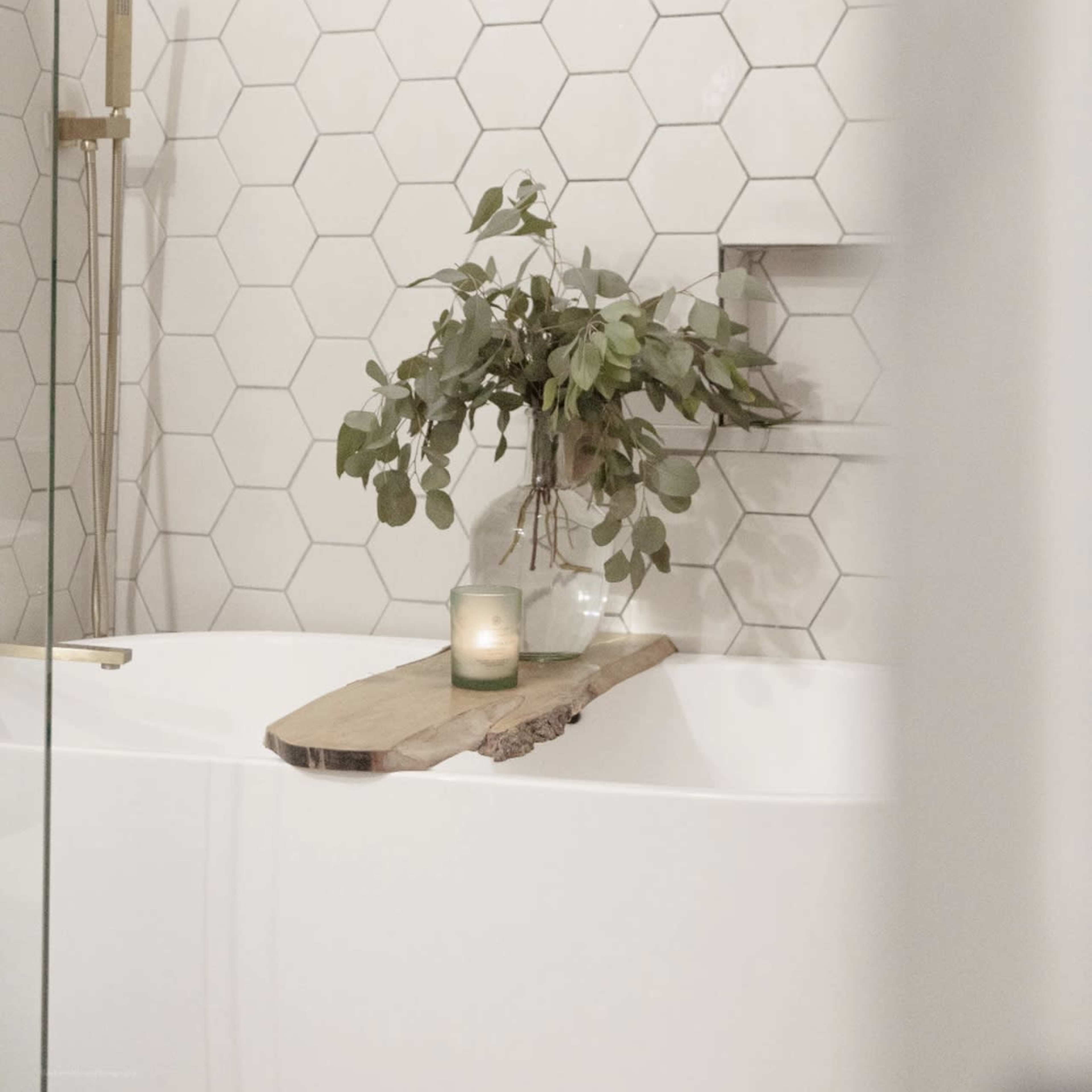 A modern bathroom features a hexagonal tile wall, a freestanding tub, and a wooden tray with a candle and a vase of greenery.