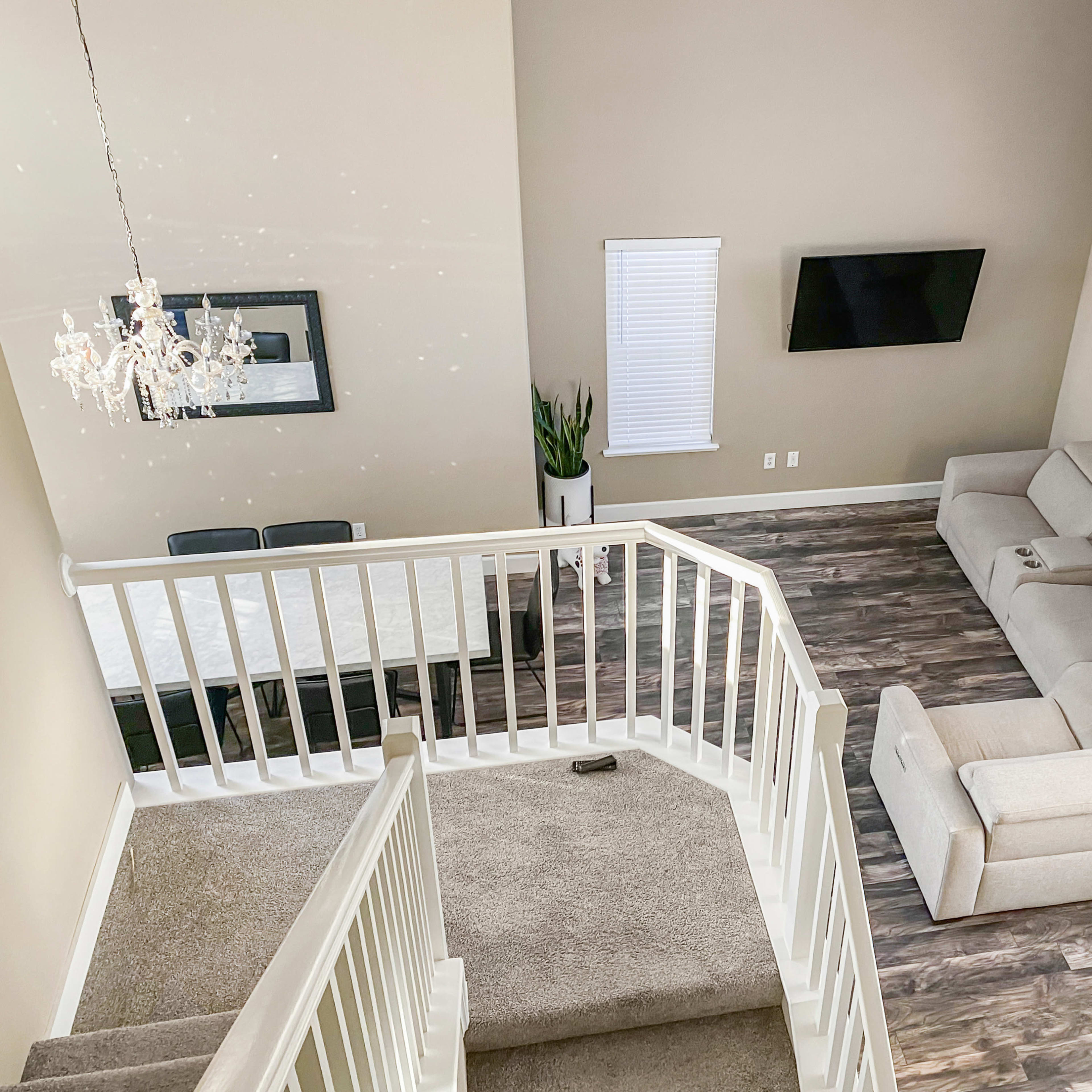 A spacious living room features a light-colored sectional sofa, a flat-screen TV mounted on the wall, and a dining area with a table, all viewed from a balcony overlooking the scene.