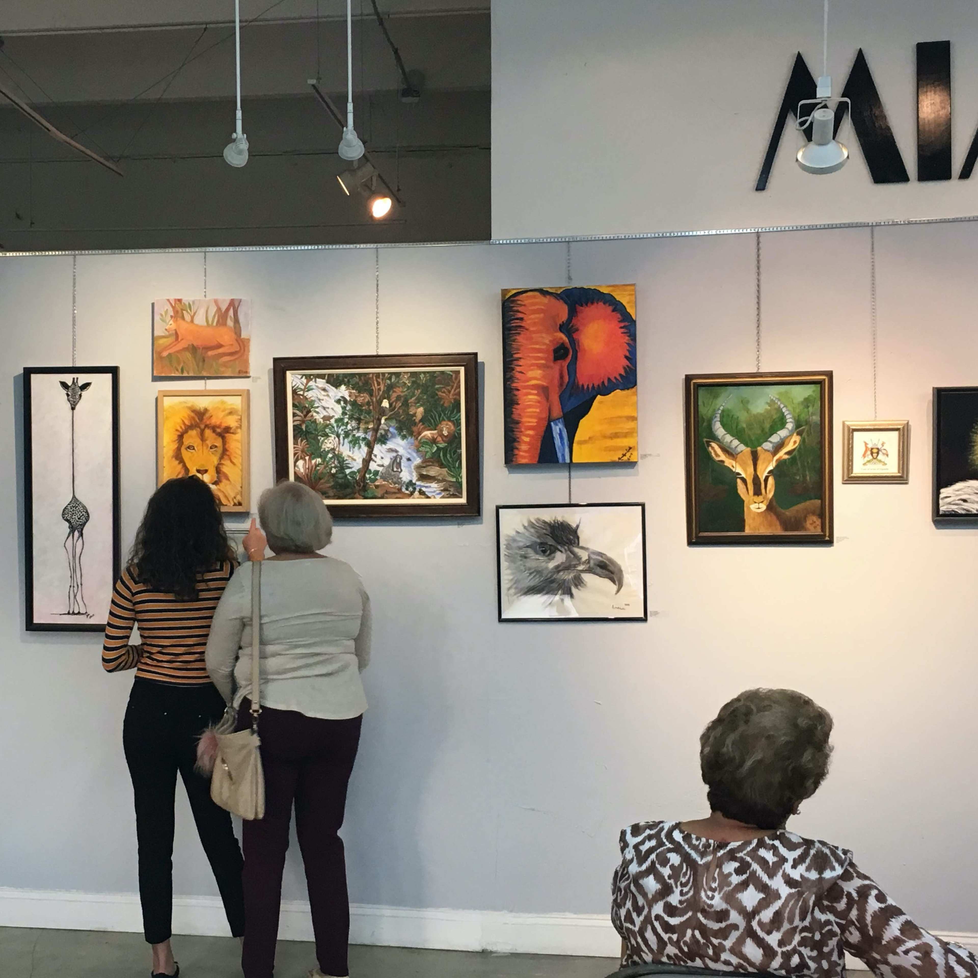 Two women observe a collection of framed artwork displayed on a gallery wall, while another woman sits in the foreground.