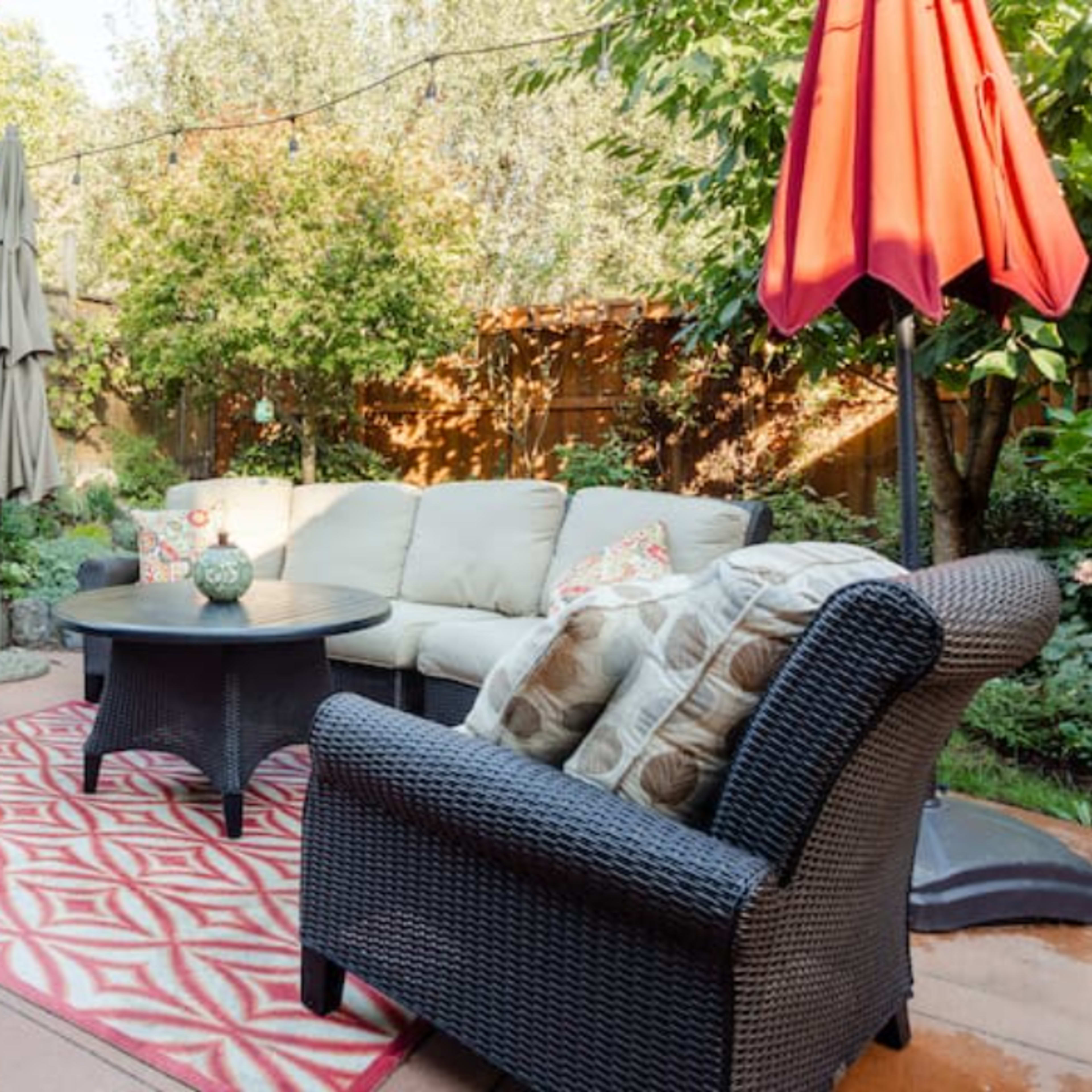 A backyard seating area with a sectional sofa, two armchairs, a round table, and a red umbrella, surrounded by greenery.