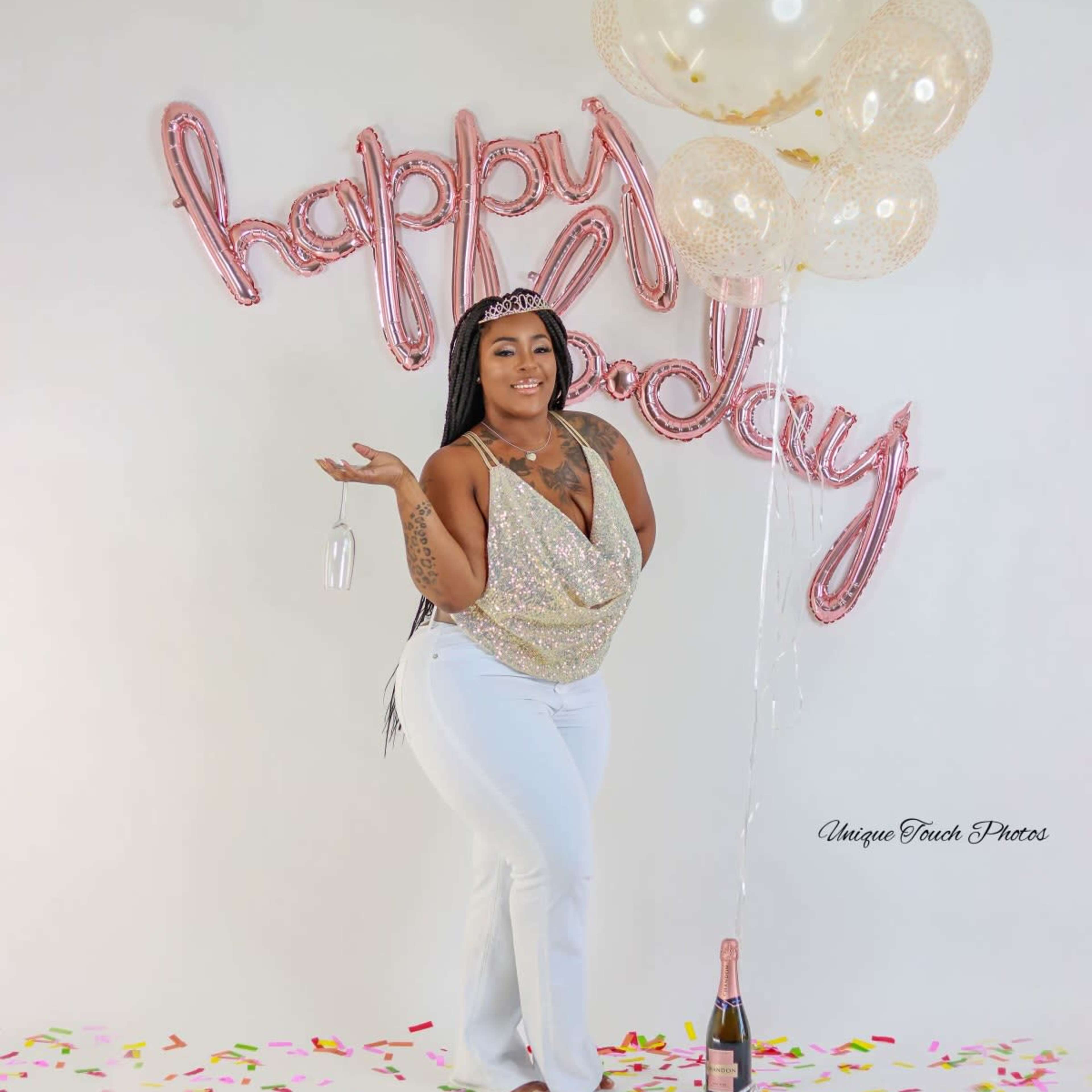 A person stands in front of a "happy birthday" balloon display, holding a champagne bottle and surrounded by colorful confetti and balloons.
