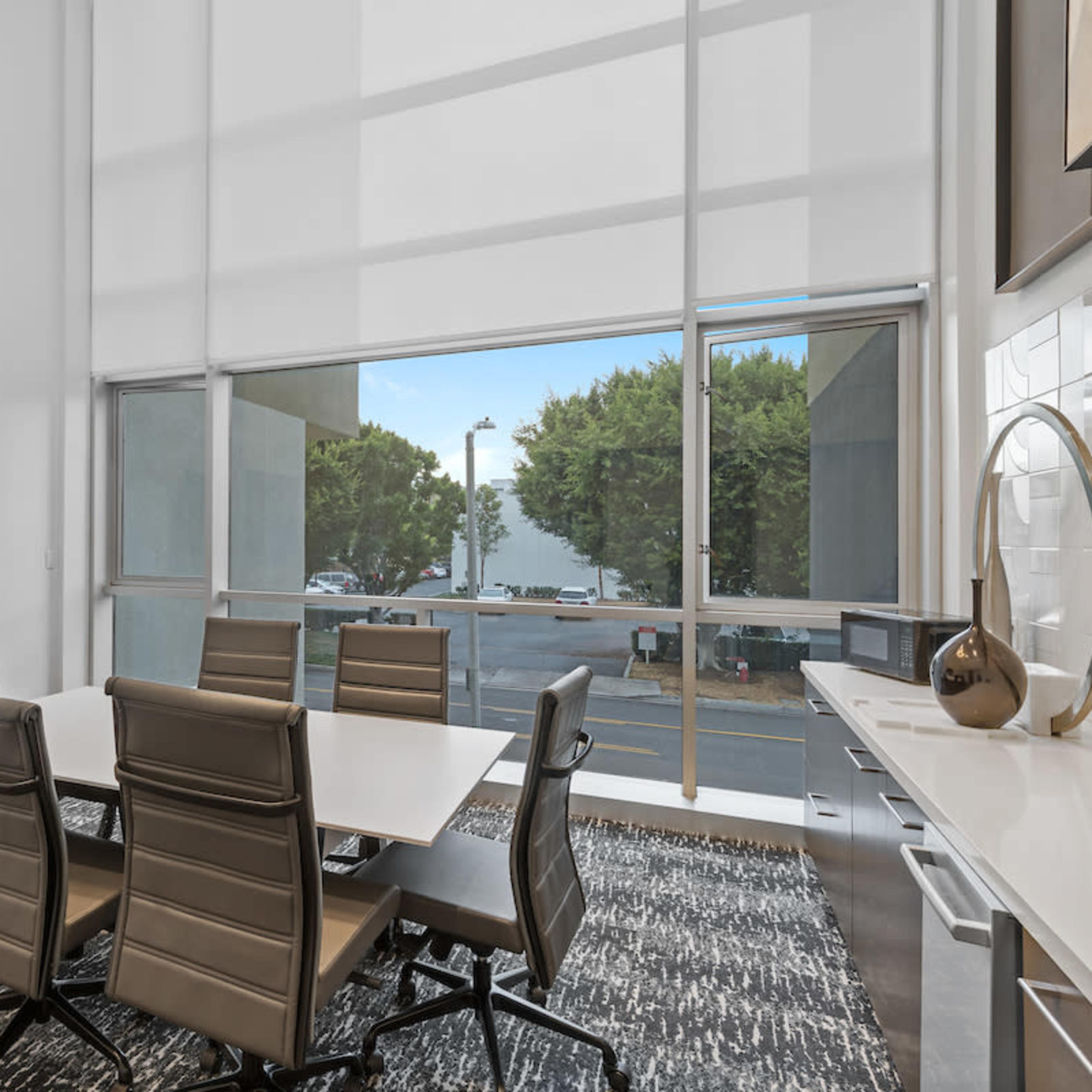 A modern conference room features a large window, a long table surrounded by six chairs, and a wall-mounted television.
