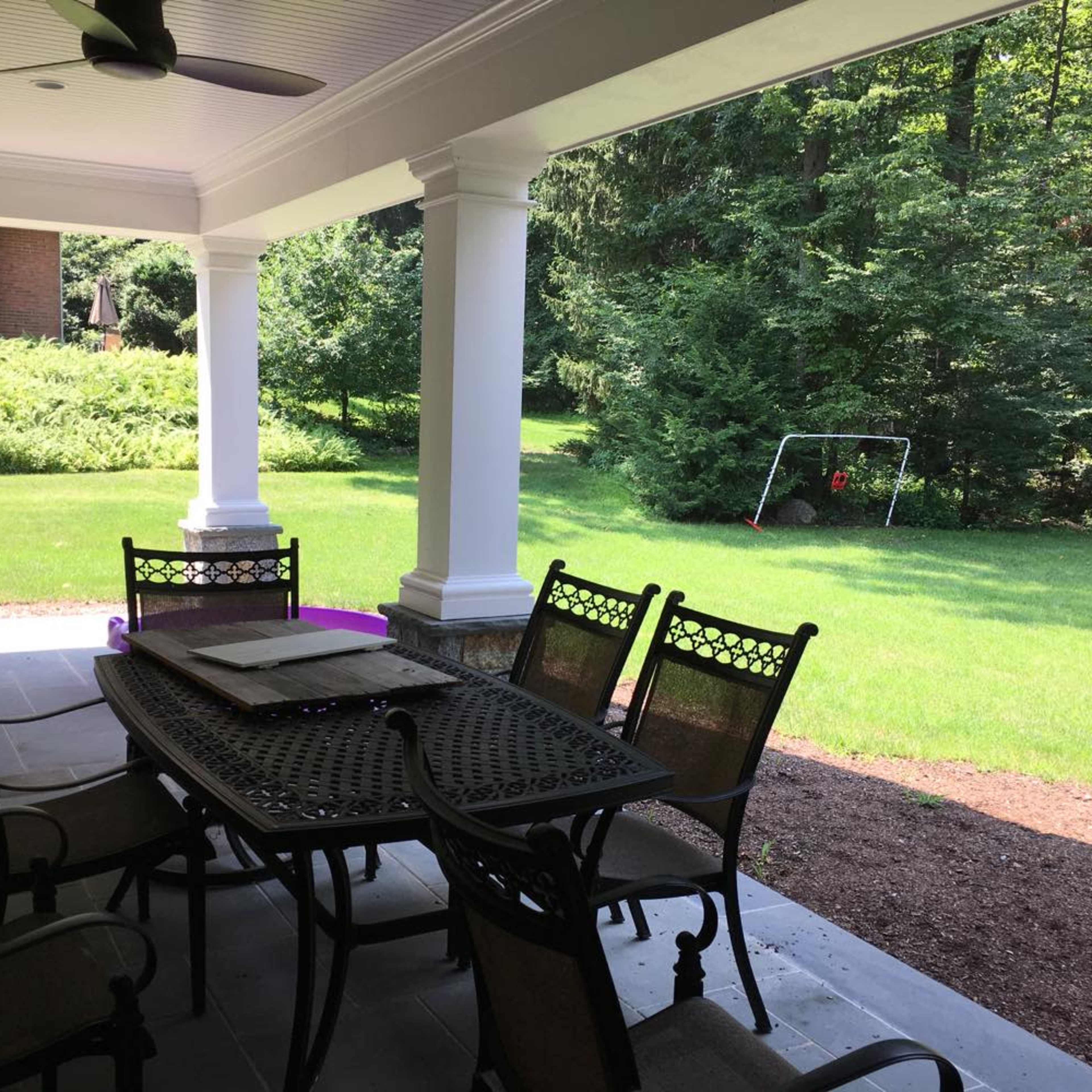 A covered patio with a dining table and chairs overlooks a green lawn and trees, with a swing set in the distance.