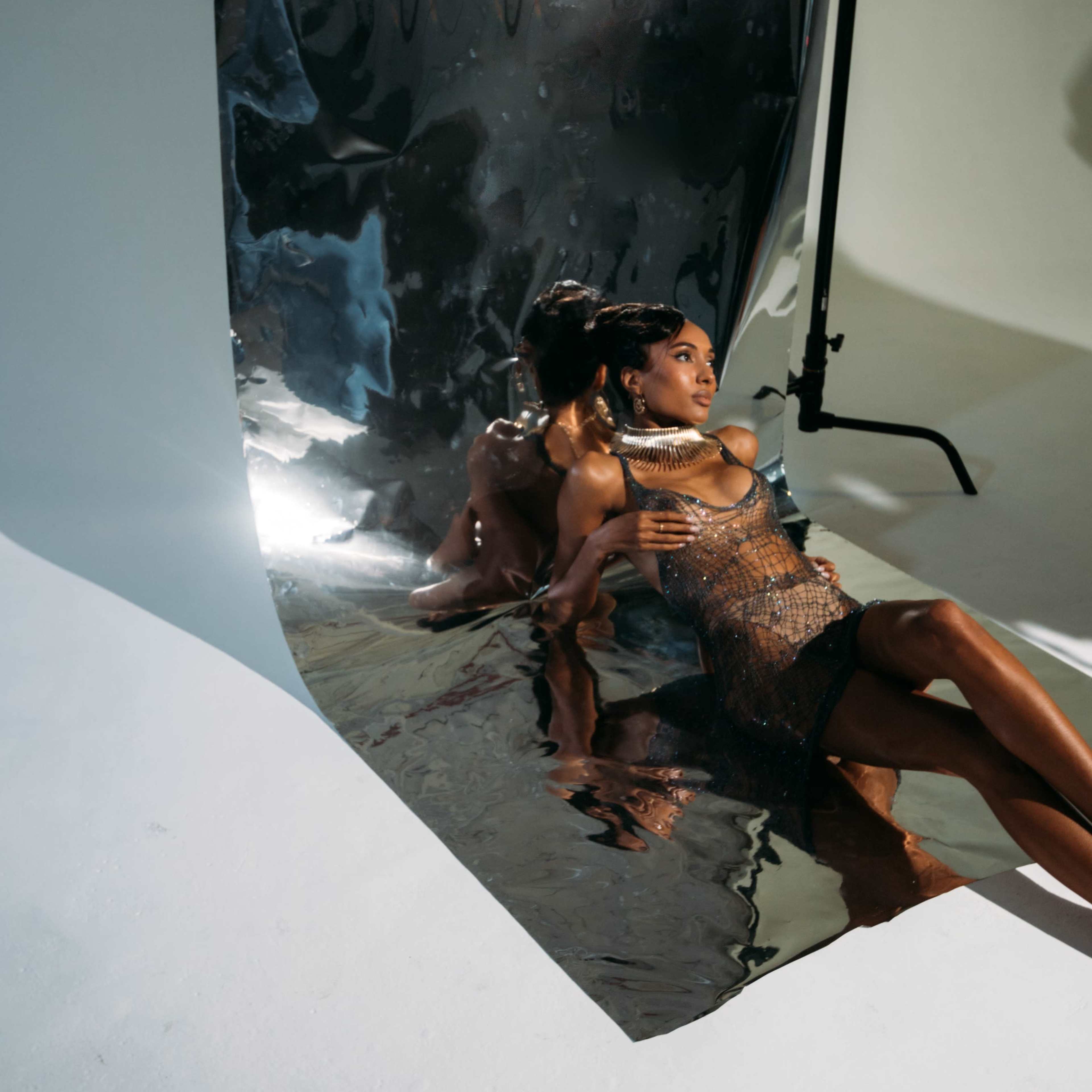 A model posed on a white surface beside a large piece of shiny, reflective material, capturing her image and shadows.