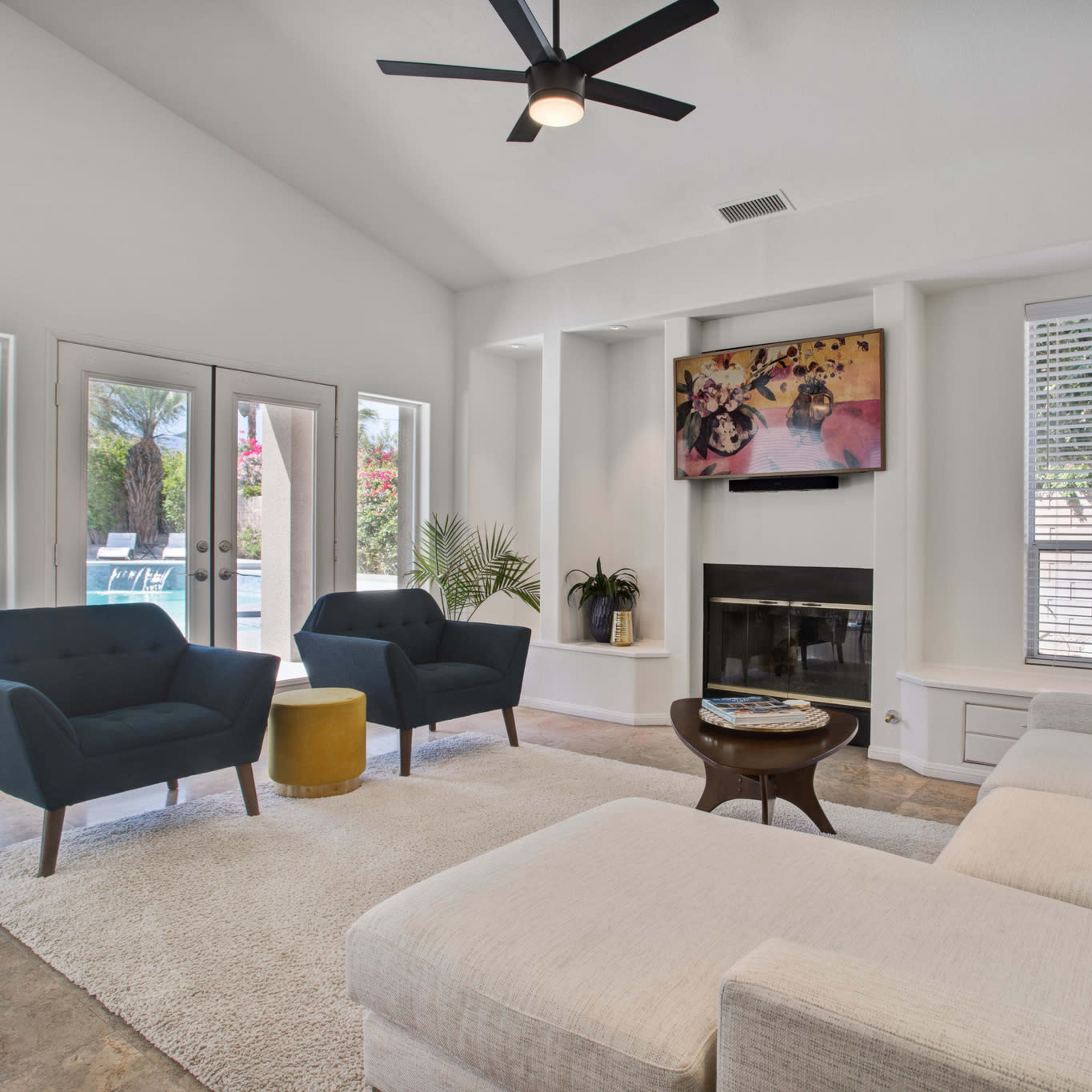 The living room features a light-colored sectional sofa, two dark blue armchairs, a round table, and large windows overlooking a pool area.