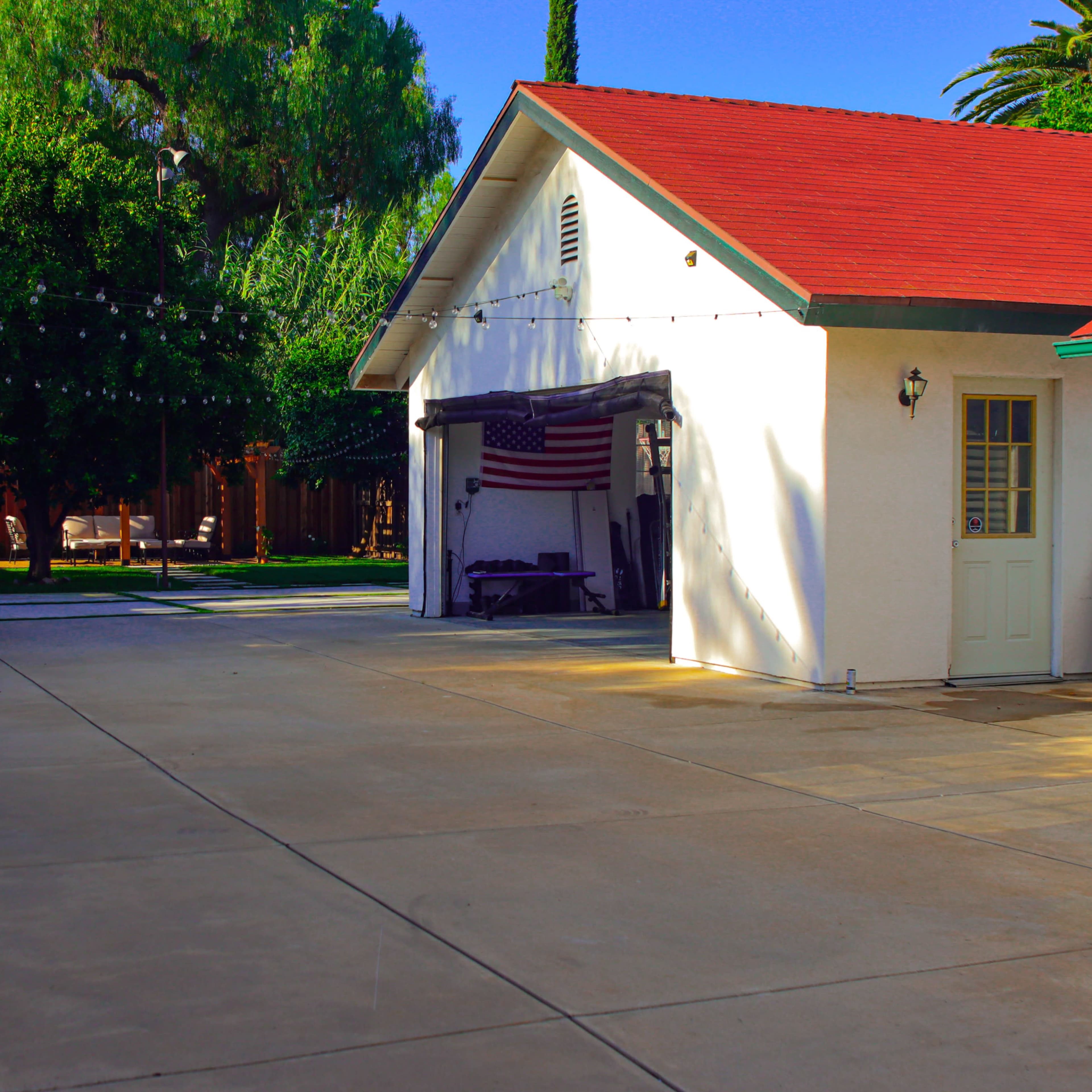 A white building with a red roof and an American flag is seen beside a clear concrete area, with green trees and outdoor seating in the background.