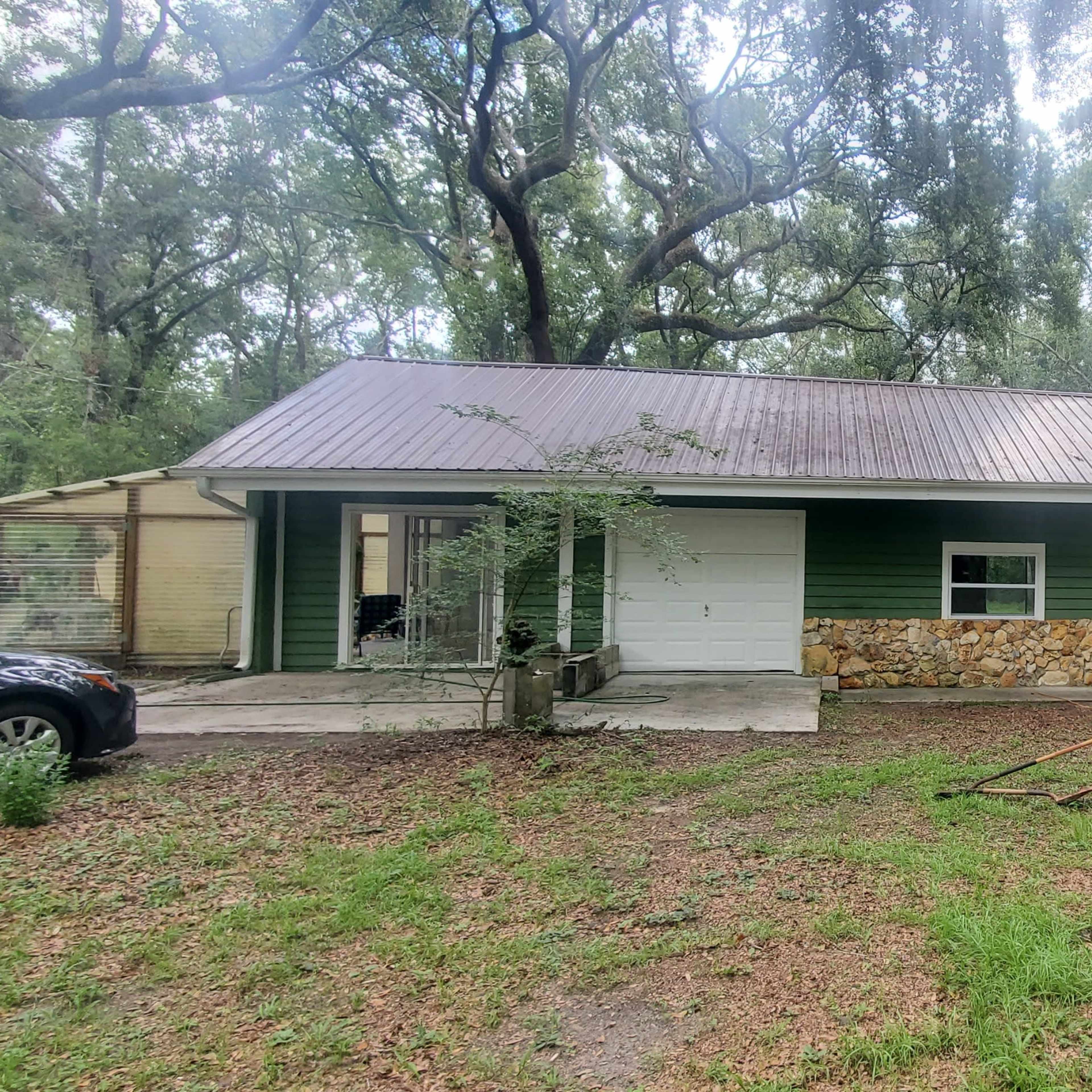 A green two-story house with a stone facade and a metal roof is surrounded by trees and a grassy area.