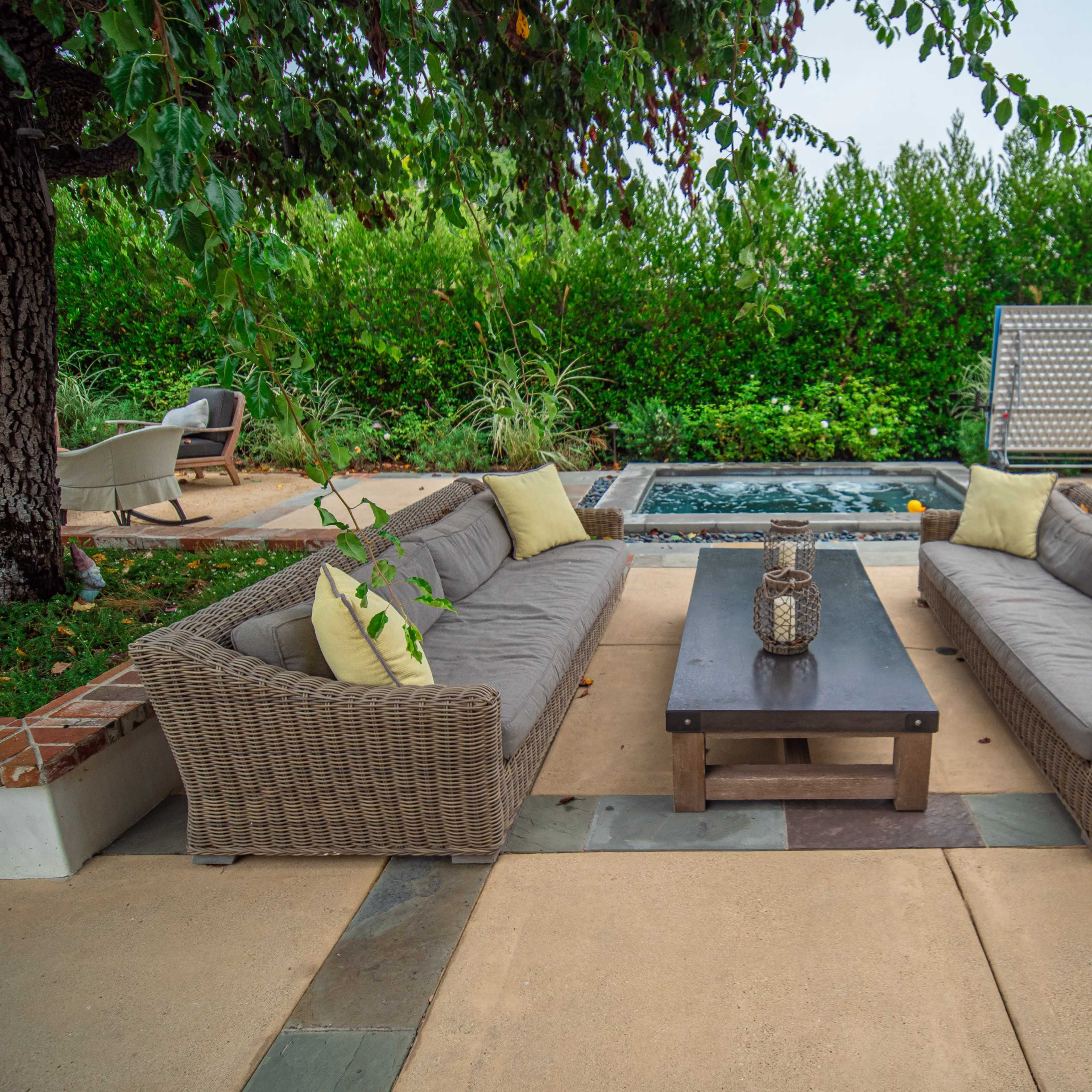 A patio area with two sectional sofas facing a coffee table, surrounded by greenery and a small pool in the background.