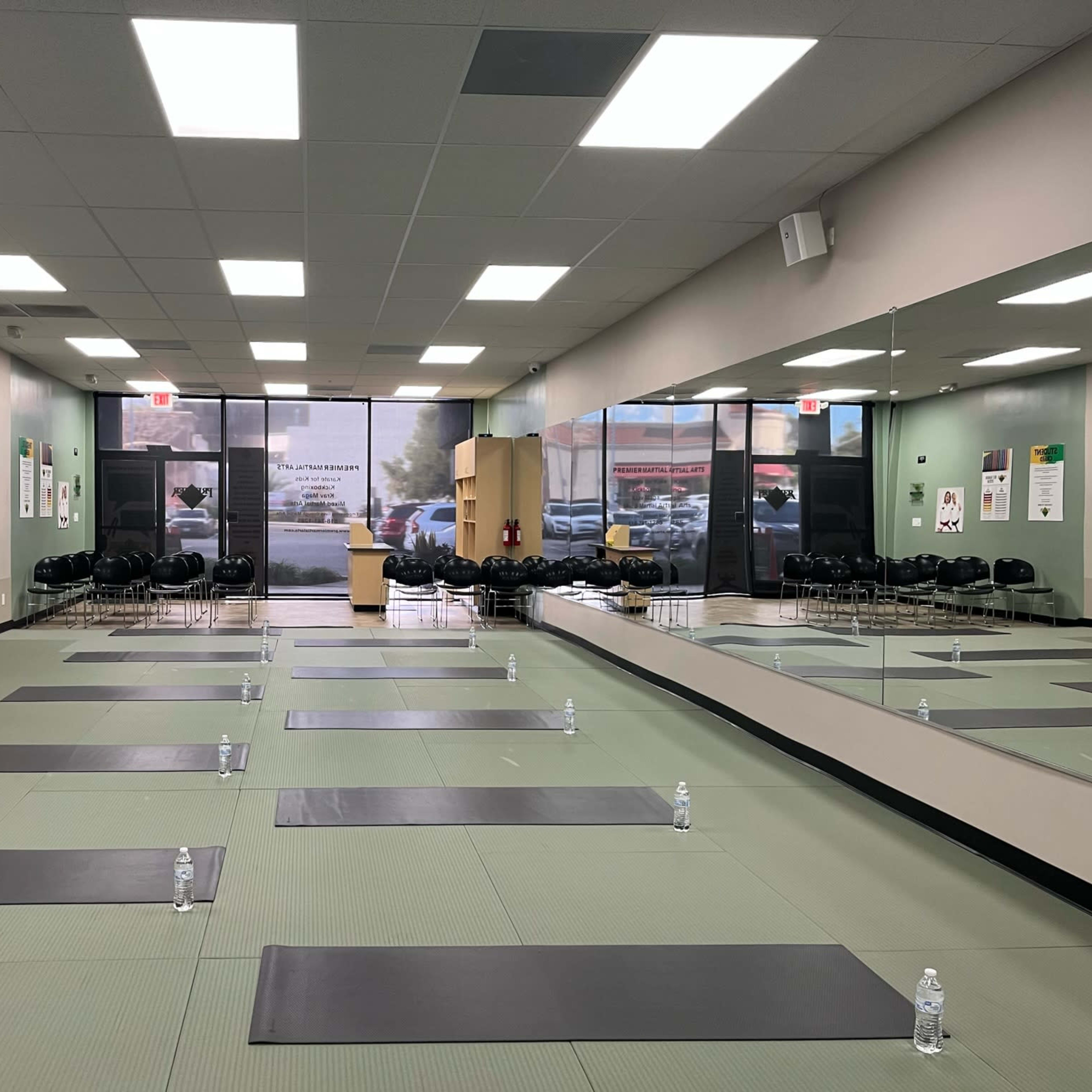 A yoga studio with several mats arranged on a green floor, water bottles placed on each mat, and a large mirror reflecting the room's interior.