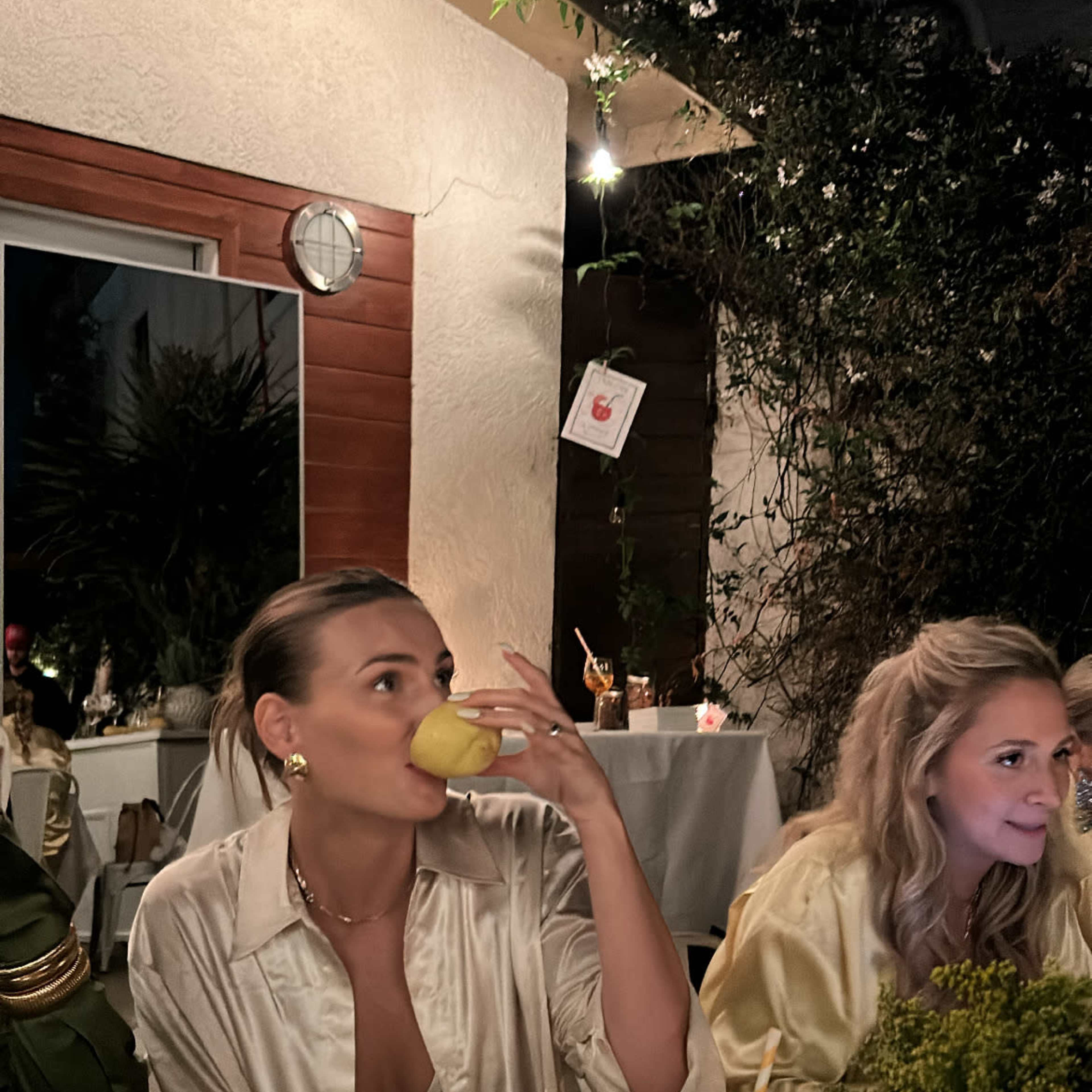 A group of women in gold outfits gathers around a decorated outdoor dining table, surrounded by greenery and modern architecture.