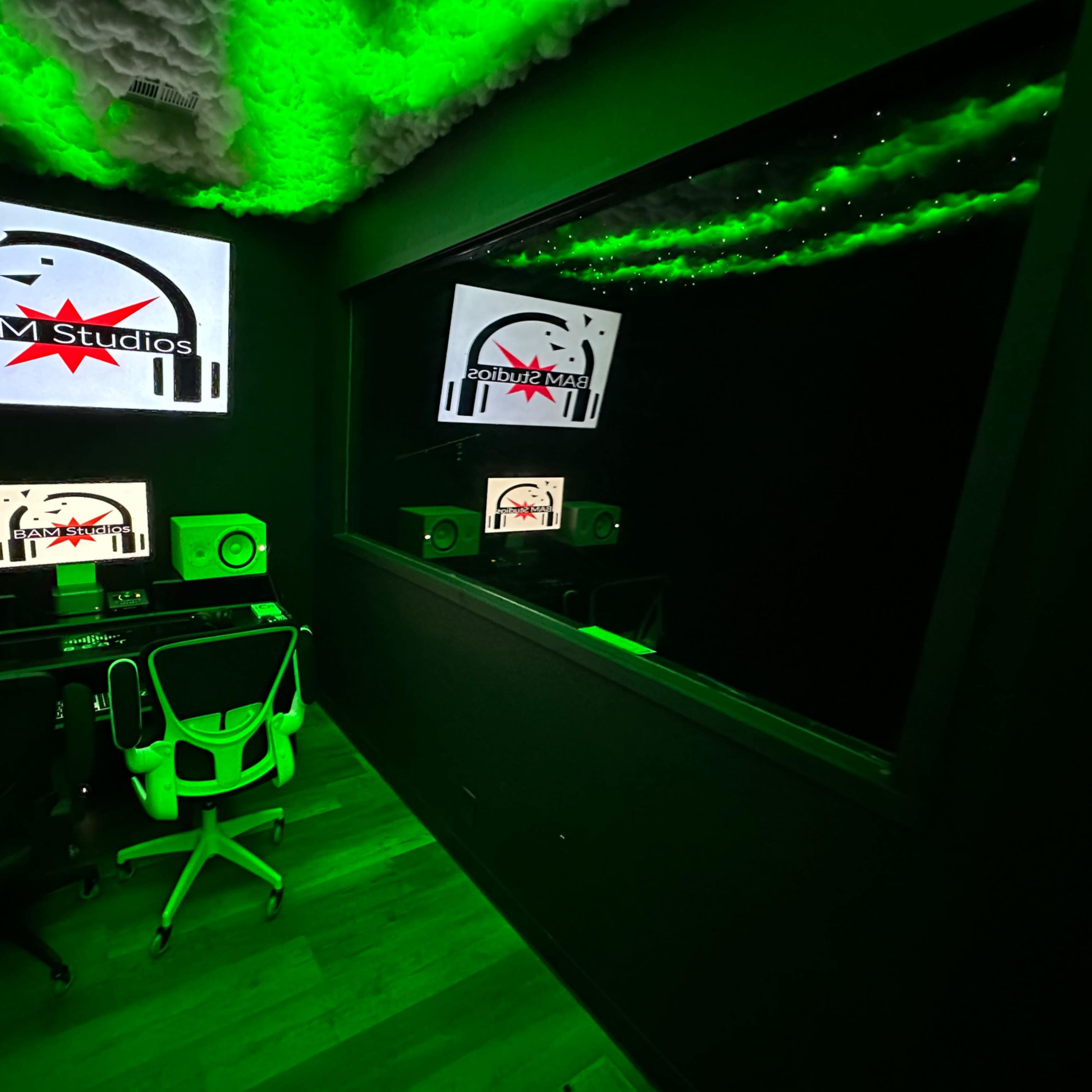 A sound studio equipped with monitors, speakers, and a large display featuring the logo of BAM Studios, with green lighting and a reflective surface.